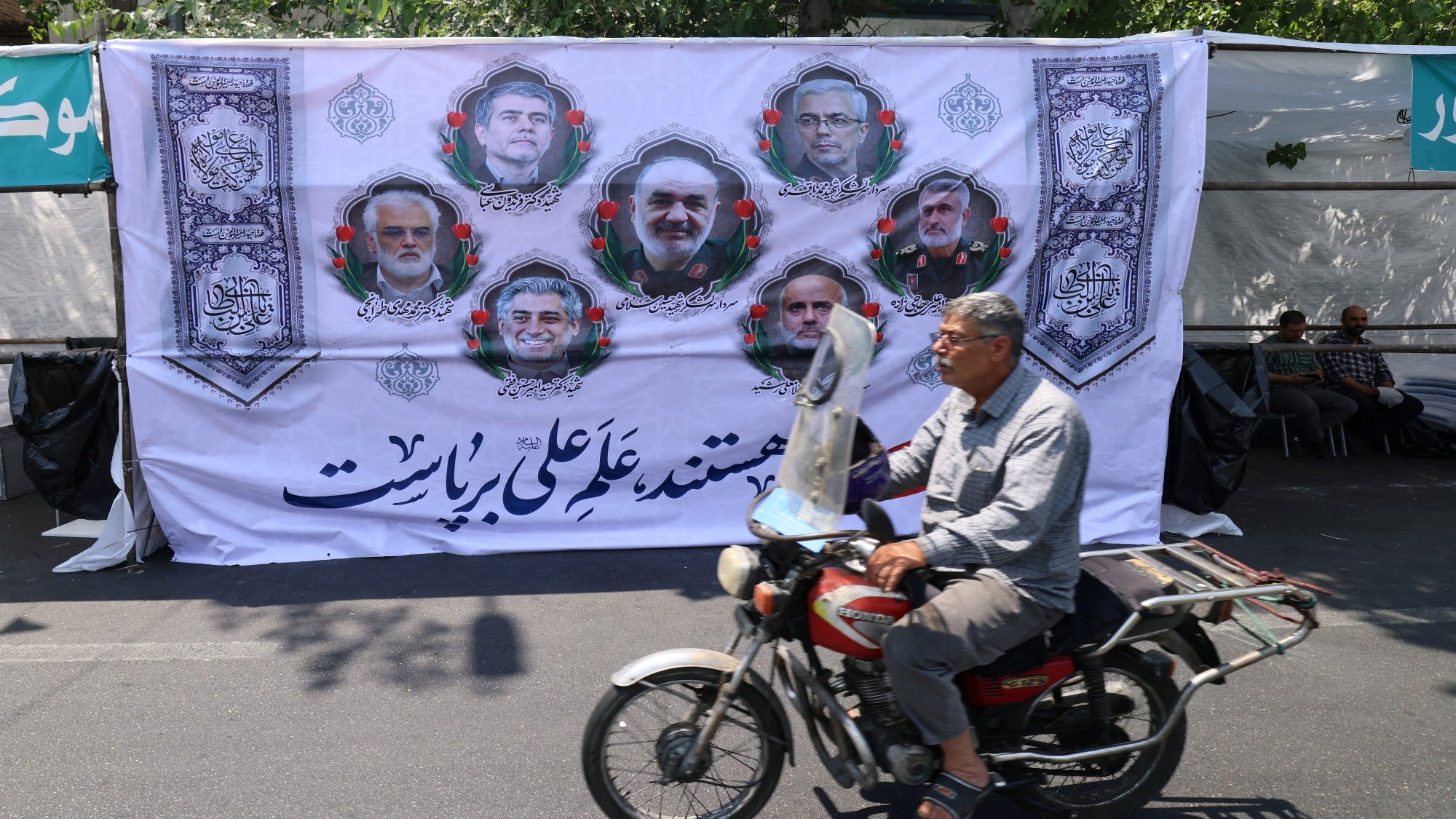 Un homme à moto passe devant une grande affiche avec des portraits et des motifs décoratifs.