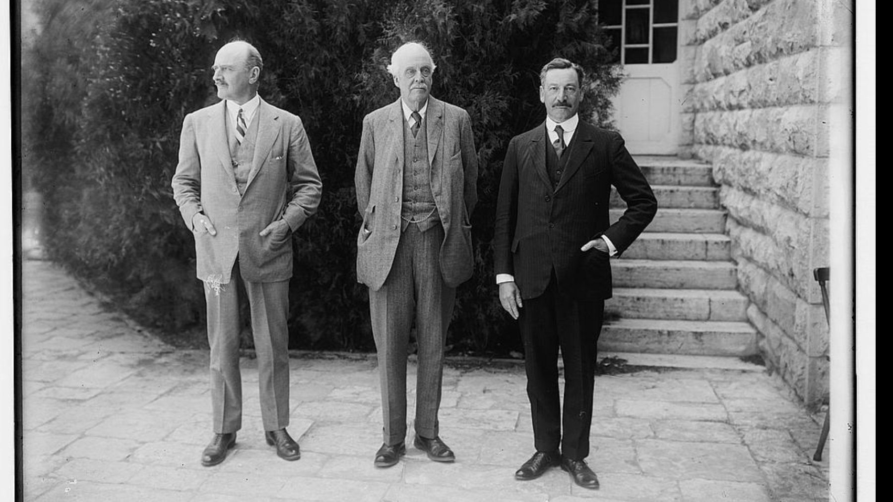 Trois hommes en costumes, debout devant un bâtiment avec des marches et des arbustes.