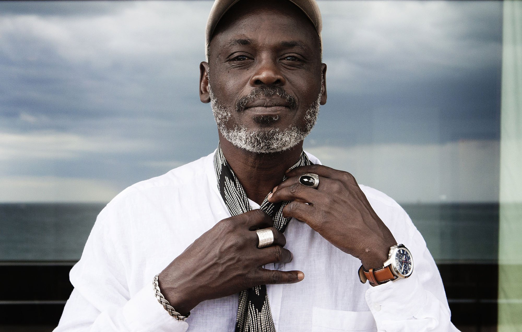 L'immagine ritrae un uomo che indossa una camicia bianca e un cappellino. Ha una barba grigia e un'espressione serena. È circondato da un ambiente luminoso, con una vista sul mare sullo sfondo. L'uomo tiene una sciarpa in mano, mentre i suoi anelli e l'orologio sul polso aggiungono un tocco di stile al suo aspetto. Il cielo appare leggermente nuvoloso, creando un contrasto interessante con l'immagine complessiva.