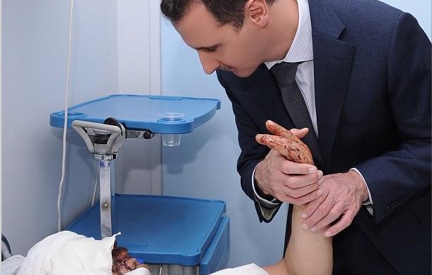 L'image montre un homme en costume qui se penche vers une personne allongée sur un lit d'hôpital, avec un bandage sur la tête. La personne blessée a une expression de souffrance, et l'homme semble lui parler ou lui apporter du soutien, tenant son bras. L'environnement est médical, avec des équipements visibles en arrière-plan.