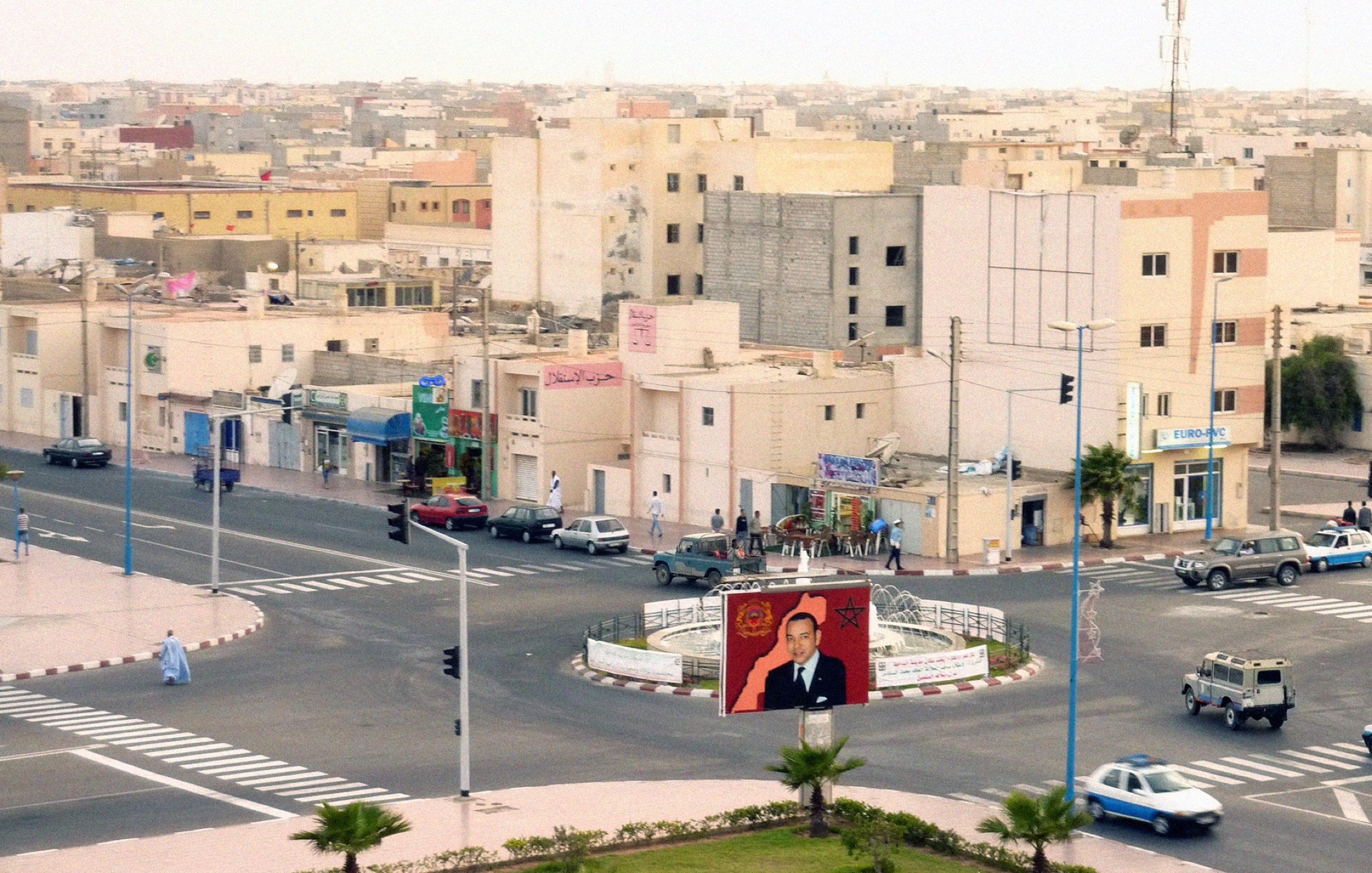 L'image montre une vue urbaine d'une ville. On y aperçoit des bâtiments de plusieurs étages, avec une architecture simple. À l'avant, il y a un rond-point avec un grand panneau affichant un portrait d'une figure publique. Plusieurs voitures sont stationnées ou circulent autour du rond-point. Des arbres et des éléments de signalisation routière ajoutent à l'ambiance de la scène. Le ciel est clair, suggérant une atmosphère calme, et la lumière semble indiquer une période de la journée avancée.