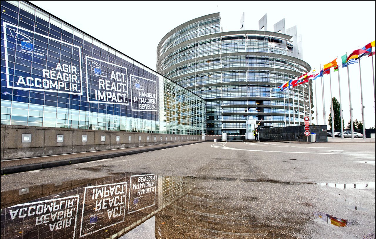 L'image représente un bâtiment moderne avec une architecture distinctive, probablement un siège d'institution européenne. On peut voir des fenêtres en verre réfléchissant des phrases comme "AGIR, RÉAGIR, ACCOMPLIR" en plusieurs langues. À droite, il y a une série de drapeaux représentant différents pays, tandis qu'une flaque d'eau au premier plan reflète l'édifice et les messages affichés sur la façade. L'ensemble donne une impression de dynamisme et de modernité.