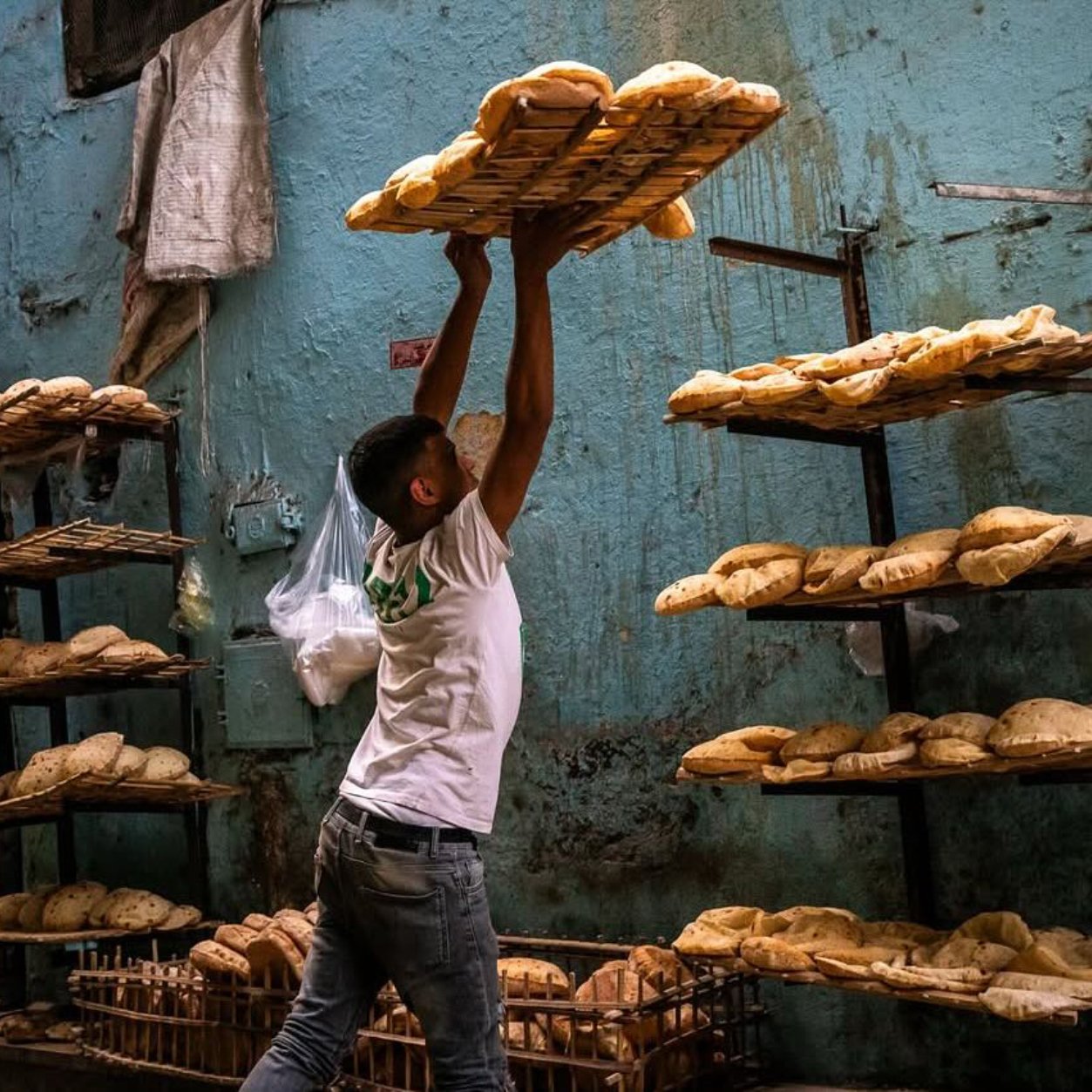 Un hombre levanta canastas con pan en un ambiente de panadería. Las paredes son azules y desgastadas.