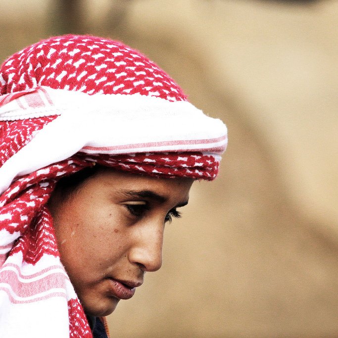 در این تصویر، یک فرد با چهره‌ای متعلق به سن جوانی دیده می‌شود. او یک کلاهی با طرح قرمز و سفید بر سر دارد که به شیوه‌ای سنتی پوشیده شده است. چهره‌اش به سمت پایین متمایل است و نگاهی تفکرآمیز دارد. پس‌زمینه تصویر مبهم است و توجه بیشتر بر روی شخص متمرکز شده است. این تصویر حس آرامش و تمرکز را منتقل می‌کند.