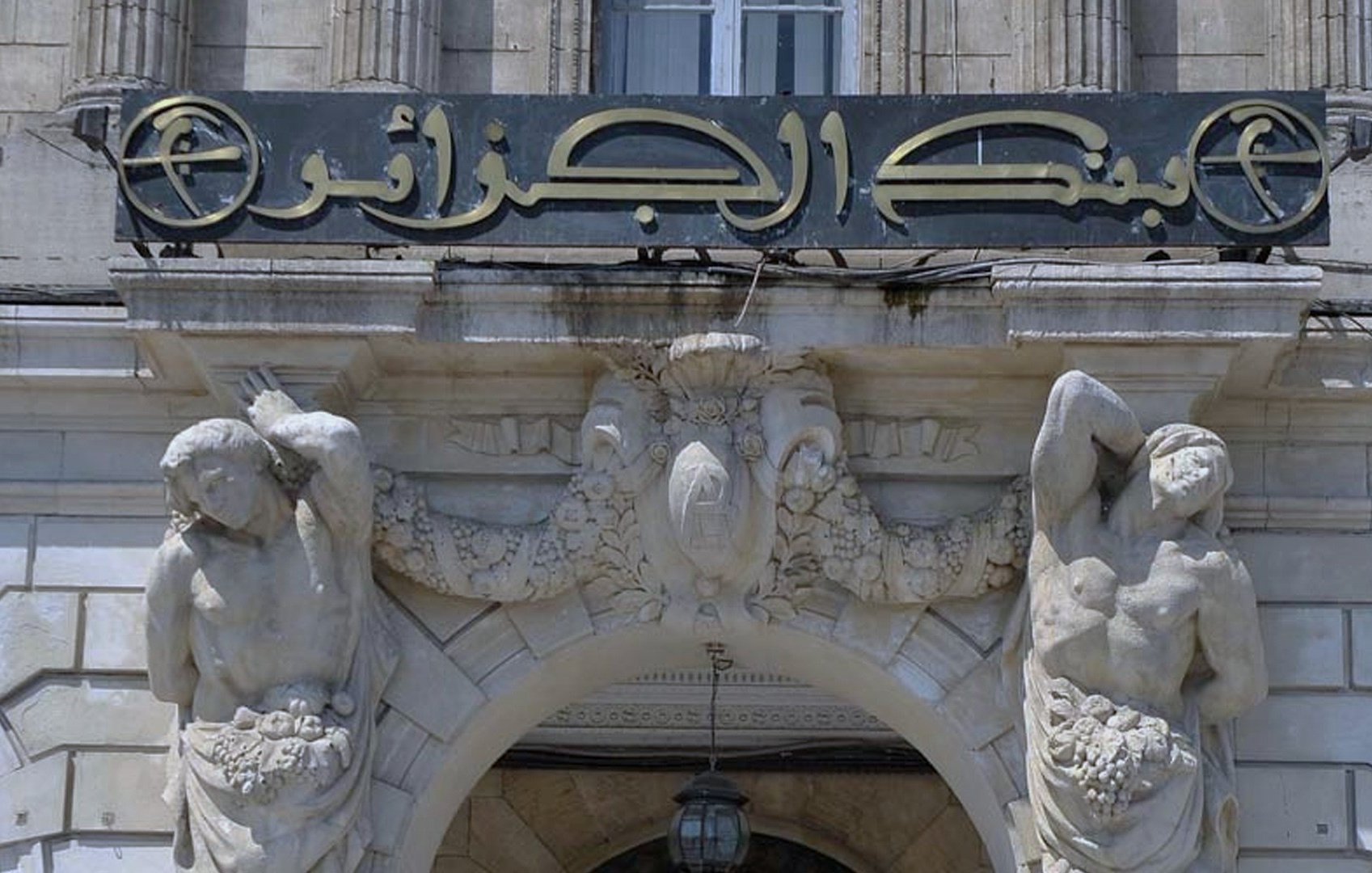L'image montre une façade architecturale ornée d'une grande enseigne avec des inscriptions en arabe. Au-dessus de l'entrée, deux sculptures de figures humaines sont placées de part et d'autre, tenant chacune des éléments décoratifs comme des grappes de fruits. L'ensemble dégage un style classique, mettant en avant des détails sculptés sur la pierre et une embellissement artistique de l'architecture.