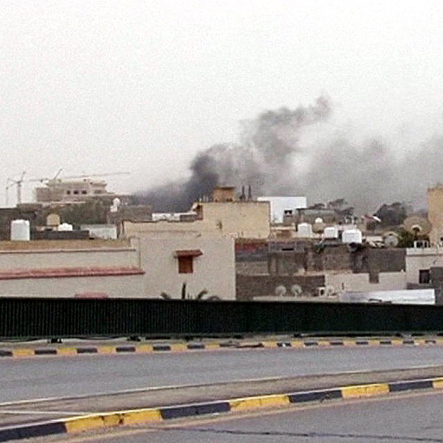 L'image montre une scène urbaine avec des bâtiments de différentes tailles, typiques d'un environnement urbain. Au loin, on peut apercevoir des colonnes de fumée sombre s'élevant dans le ciel, suggérant probablement un incendie ou une explosion. Le ciel est nuageux, ce qui ajoute à l'atmosphère sombre de la scène. Au premier plan, une personne marche le long d'une route, semblant s'éloigner de la source de la fumée. Les couleurs sont plutôt ternes, reflétant une ambiance de tension ou d'urgence.
