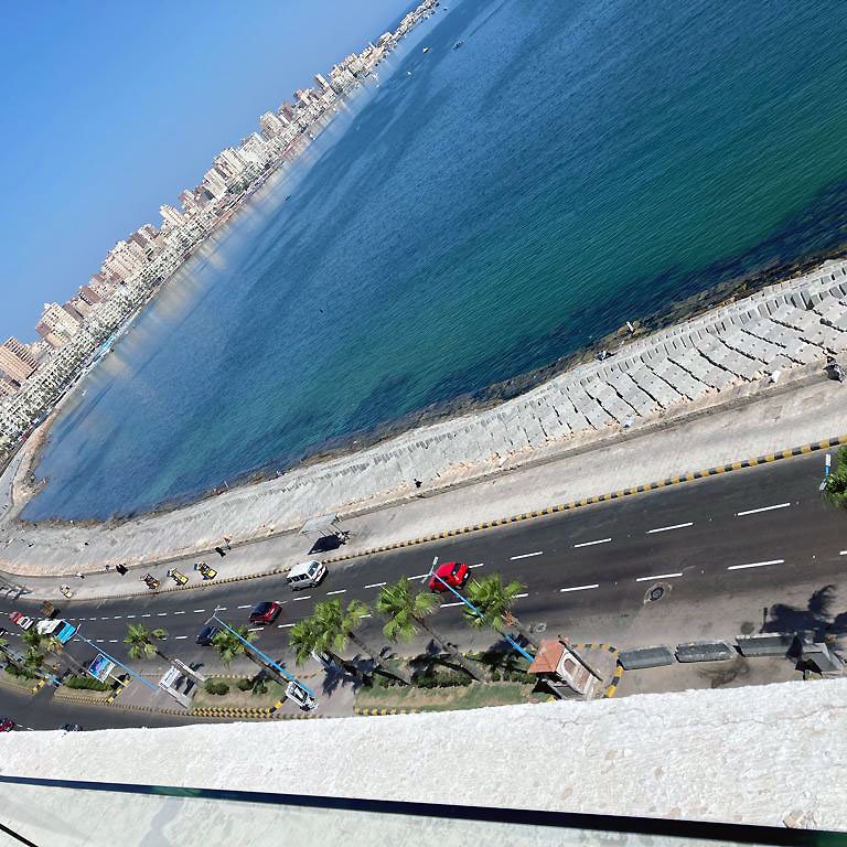 الصورة تظهر مشهداً لجانب من البحر، حيث يمتد الشاطئ على الواجهة البحرية. يحيط بالمكان الطريق الذي يمتد على طول الساحل، ويظهر فيه عدد من الأشجار والنخيل. في الخلفية، يمكن رؤية مجموعة من المباني السكنية أو الفنادق التي تعكس نمط الحياة الحضري. لون المياه يبدو زرقاً صافياً، مما يضيف جمالاً إلى المشهد.