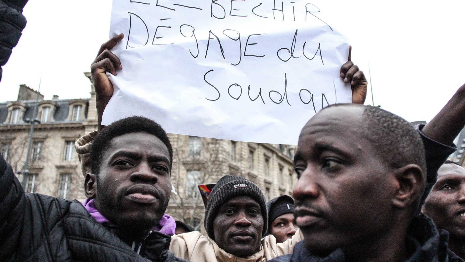 L'image montre une manifestation où des personnes tiennent des pancartes. L'une d'elles est clairement visible et affiche un message en français disant "El-Béchir dégage du Soudan". Les participants semblent passionnés et engagés dans une cause. On peut discerner des expressions sérieuses et déterminées sur leurs visages. En arrière-plan, des bâtiments peuvent être aperçus, suggérant un cadre urbain.
