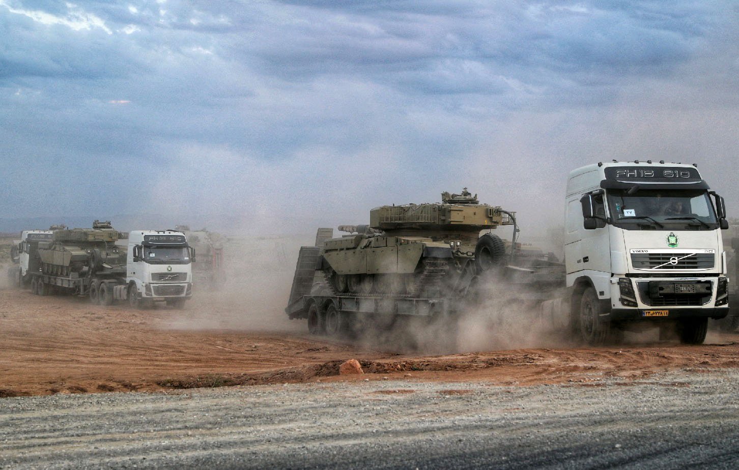 L'image montre une scène de transport militaire, avec plusieurs camions, notamment un portant le logo Volvo, transportant des véhicules blindés. La poussière s'élève autour des véhicules, suggérant un environnement saharien ou aride. Le ciel est nuageux, ajoutant à l'atmosphère de la scène. Les camions avancent sur une route en terre, mettant en avant un aspect logistique lié aux opérations militaires.