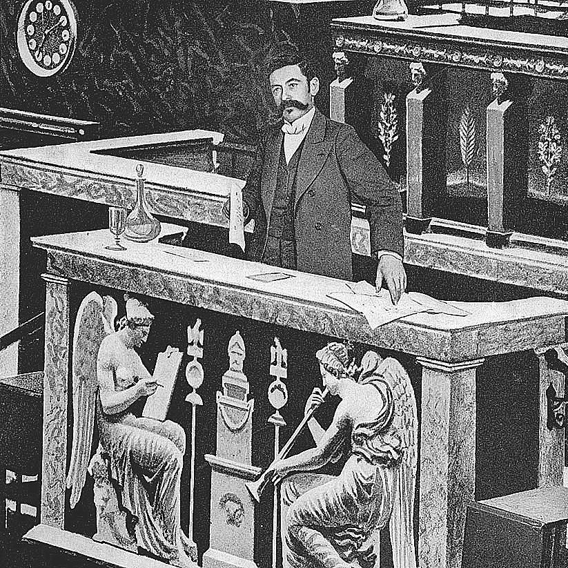 The image depicts a man standing at a podium in what appears to be a legislative or parliamentary setting. He is dressed in a formal suit and has a mustache. The podium is richly decorated, with sculptures of angelic figures and ornate designs. There seem to be documents and a glass on the podium, indicating a formal presentation or discussion taking place. The background suggests an elegant and historical architecture, typical of governmental buildings.