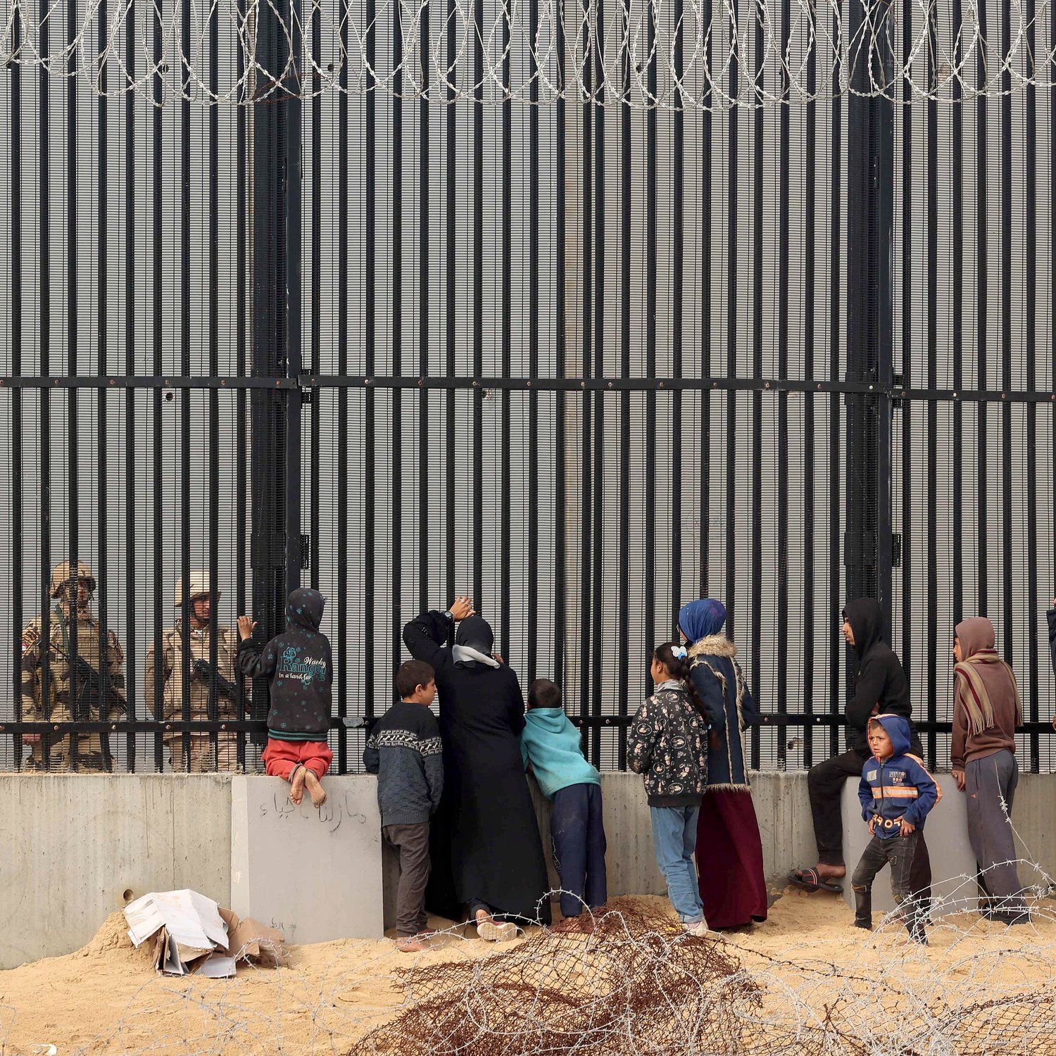 تظهر الصورة مجموعة من الأشخاص الواقفين أمام حواجز حديدية أو سياج مسنن، حيث يواجهون جنود أو عناصر أمنية على الجانب الآخر. يبدو أن هناك أطفالًا ونساء ورجالًا، وبعضهم يحملون أكياسًا أو متعلقات أخرى. البيئة المحيطة موحشة، مع وجود سلك شائك في المقدمة، مما يوضح التوتر والمشاعر القاسية في هذا المشهد.