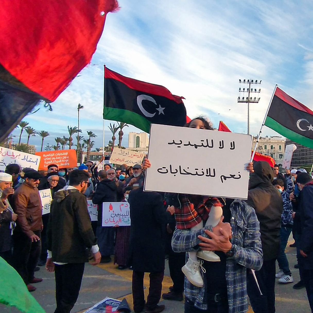 تظهر الصورة مشهداً من تظاهرة في مكان عام، حيث يحتشد الناس حاملين لافتات تتضمن عبارات تتعلق بالحرية والانتخابات. في الخلفية، ترفرف الأعلام الليبية بألوانها الأسود والأحمر والأخضر، مما يدل على الهوية الوطنية. يبدو أن الحشد يتنوع بين الرجال والنساء، وبعضهم يرتدي الكمامات، مما يشير إلى الحرص على الصحة العامة. السماء في الخلفية تظهر مع تزاحم الغيوم، مما يضيف جواً مميزاً إلى البيئة المحيطة بالتجمع.