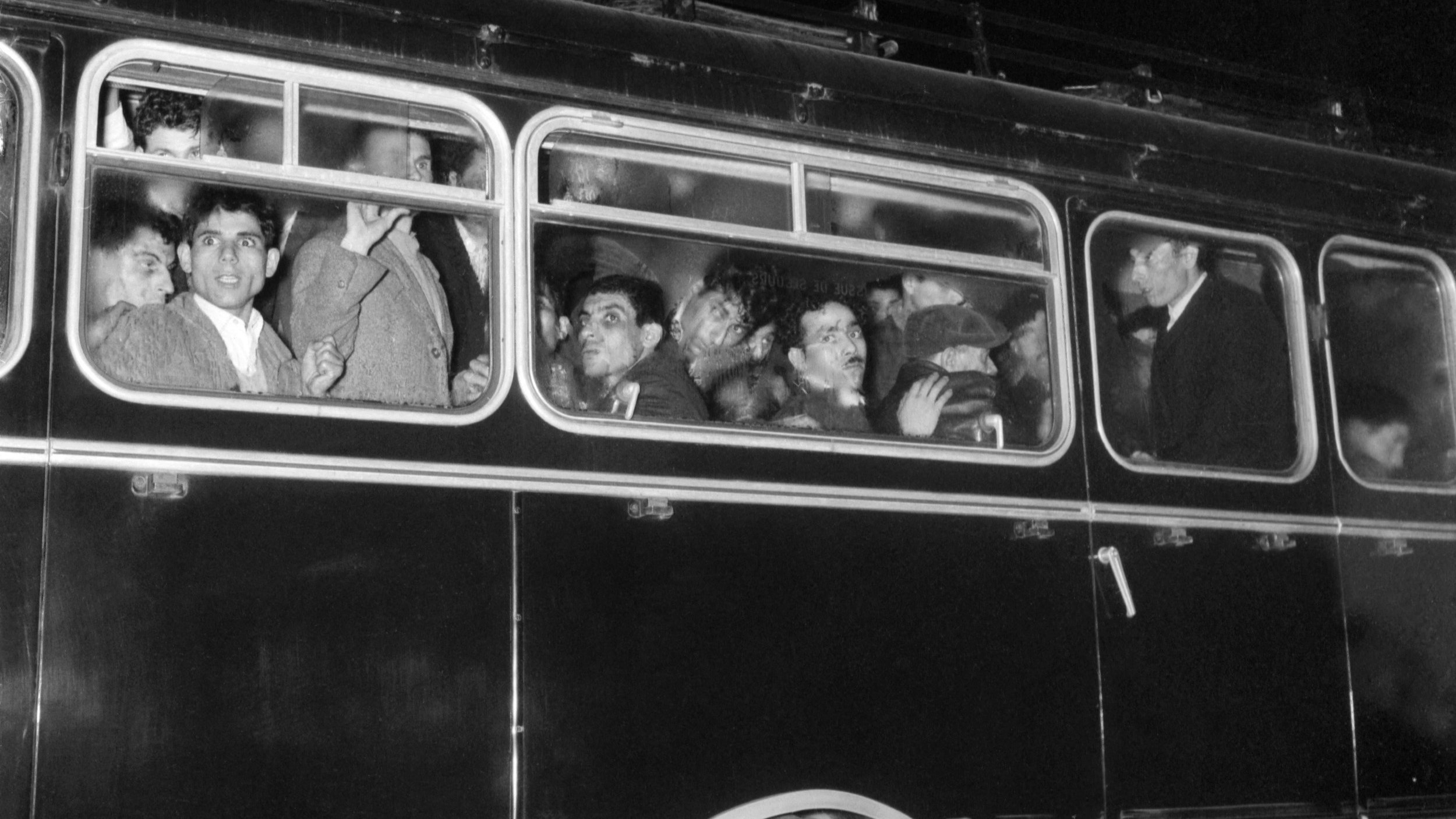 The image depicts a crowded bus at night. Several people are visible through the windows, some looking out while others appear to be engaged in conversation. The bus is dark, possibly painted black, and has a classic design with multiple windows. The expressions of the individuals range from curious to contemplative, highlighting a moment of shared experience among the passengers. The scene captures a sense of movement and typical urban life.