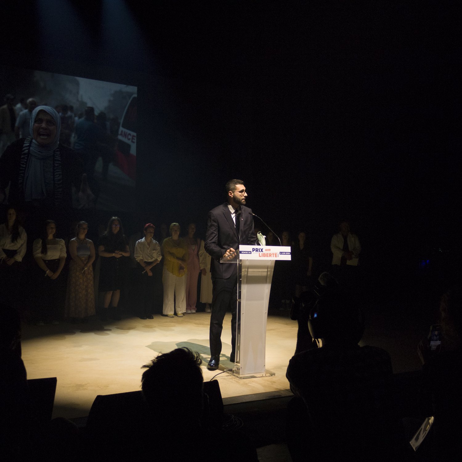 L'immagine mostra un uomo che parla al microfono su un palco, mentre sullo sfondo si vede un'immagine proiettata di una donna con un velo e altre persone. L'oratore indossa un abito elegante e sembra essere il centro dell'attenzione. Dietro di lui, altre persone sono schierate, probabilmente in segno di supporto o partecipazione all'evento. L'illuminazione è drammatica, creando un'atmosfera intensa e concentrata sull'oratore.