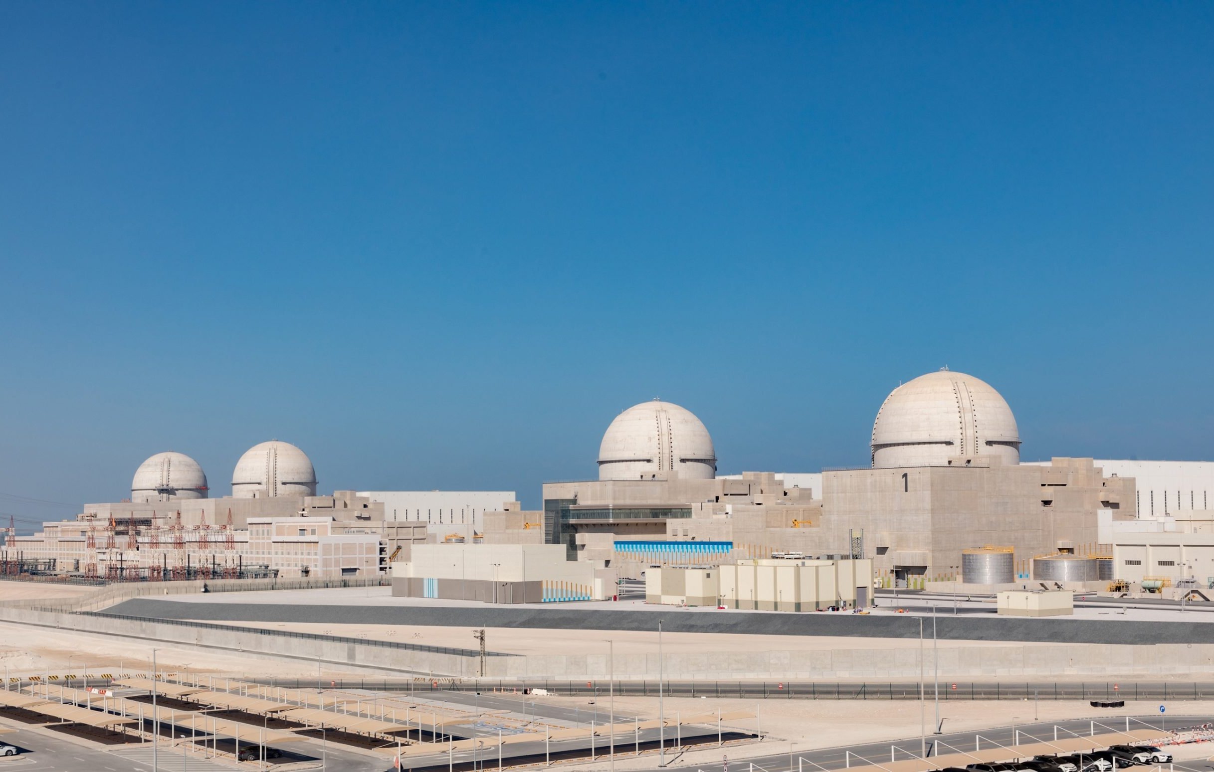 The image shows a large industrial facility, likely a nuclear power plant, with several prominent dome-shaped structures. The plant features a modern design with various buildings and infrastructure surrounding it, set against a clear blue sky. The setting appears to be an arid or semi-arid environment, with minimal vegetation. There are parking areas and some infrastructure elements visible in the foreground, indicating it is a well-developed site.