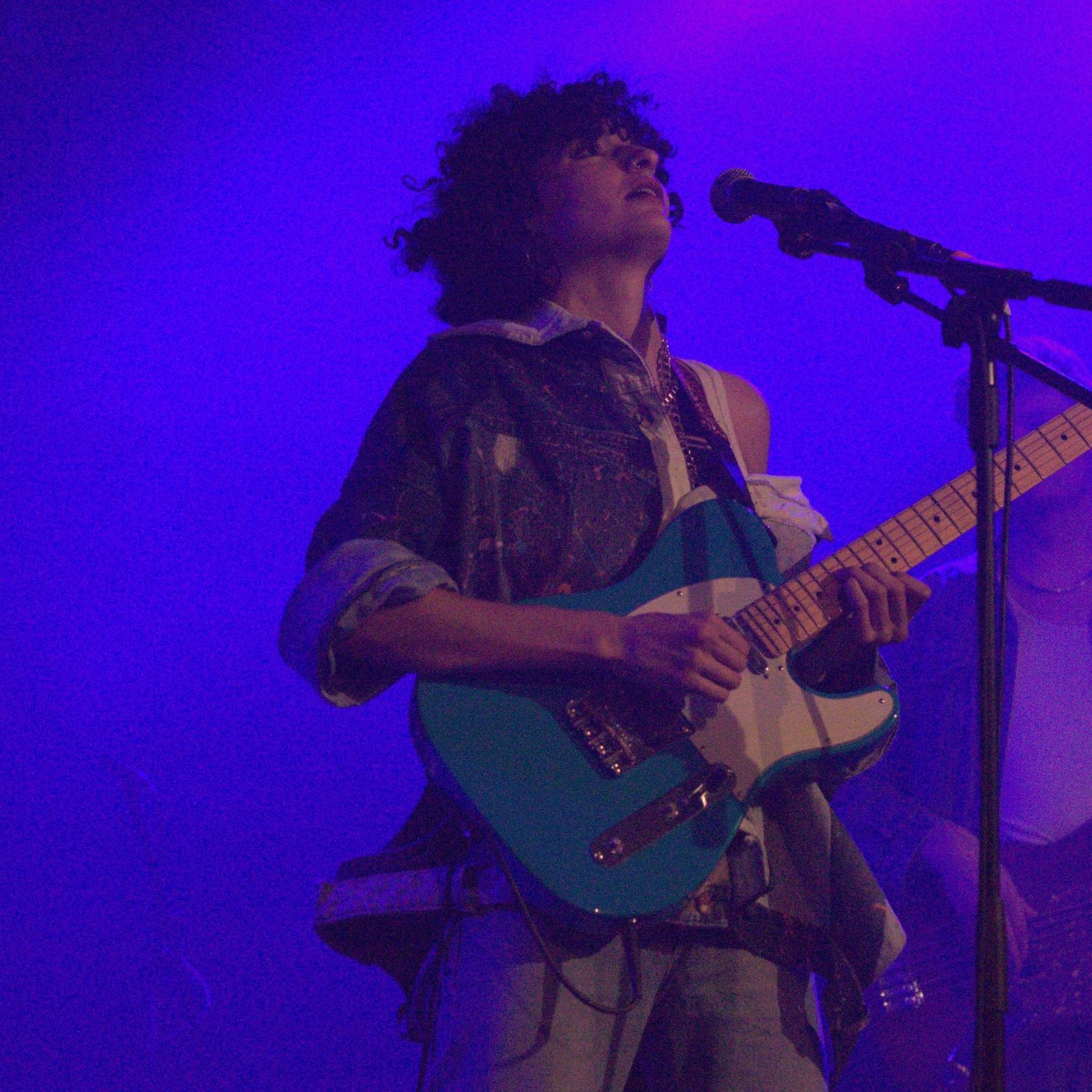 الصورة تظهر عازف جيتار على المسرح. الشخص يرتدي ملابس غير تقليدية ويعزف على آلة الجيتار الكهربائية ذات اللون الأزرق، بينما يبدو أنه يركز في الأداء. الخلفية مضيئة بلون أرجواني، مما يضيف جوًا مميزًا إلى الصورة. يمكن رؤية بعض الآلات الموسيقية الأخرى في الخلفية، مما يشير إلى وجود فرقة موسيقية.