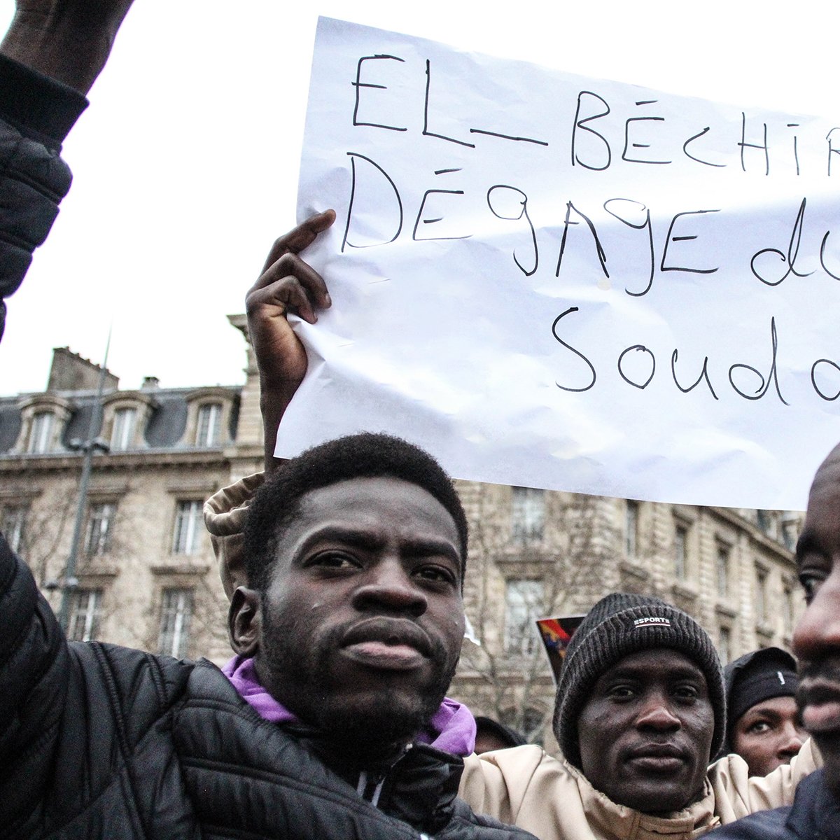 در این تصویر، تعدادی از افراد در تجمعی در حال اعتراض هستند. یک نفر در جلو، تابلویی را که روی آن نوشته شده "El Béchir dégage du Soudan" (به معنای "ال بشیر از سودان برو") را بالا گرفته است. این تجمع به نظر می‌رسد که به مسائل سیاسی و اجتماعی در سودان مربوط باشد. افراد دیگر نیز در اطراف او قرار دارند و توجهشان به تابلو جلب شده است. فضای عمومی و احساسات معترضان در تصویر به وضوح قابل مشاهده است.
