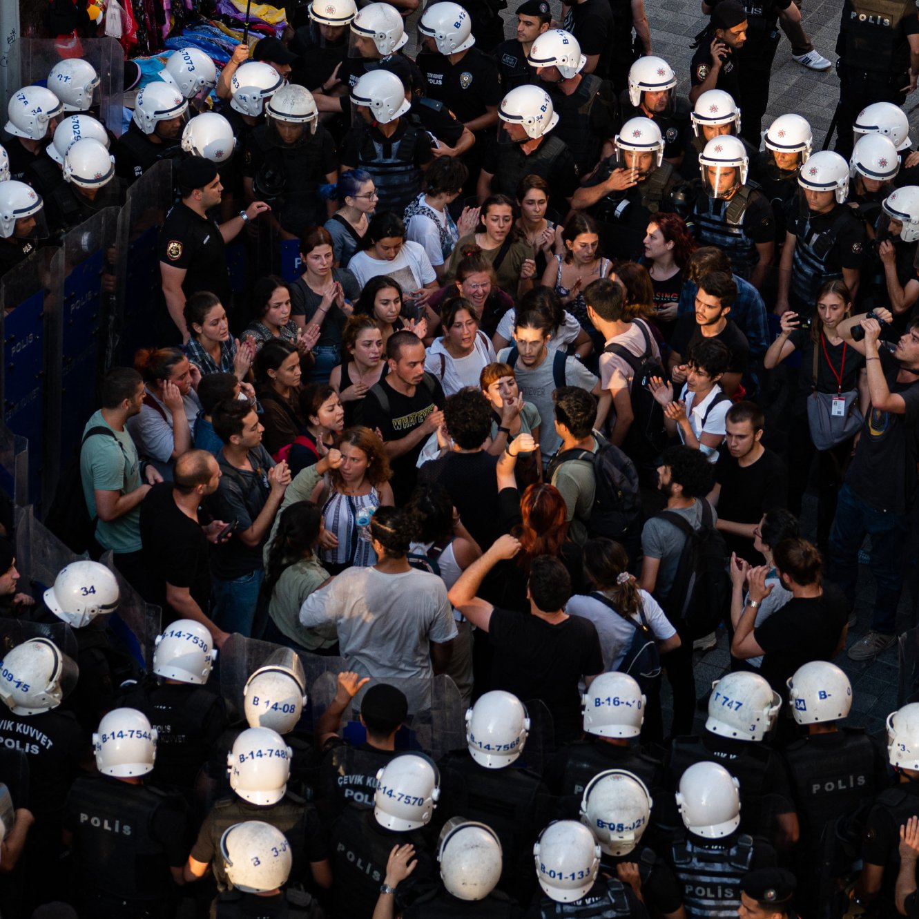 La imagen muestra una multitud de personas rodeadas por un grupo de policías. Los manifestantes parecen estar en una discusión o protesta, expresando sus opiniones entre ellos. Los policías, con cascos y uniformes, forman un cordón alrededor de la multitud, lo que sugiere una situación tensa. La escena parece tener lugar en un ambiente urbano, posiblemente durante una manifestación o conflicto social. La iluminación sugiere que puede ser al atardecer o al final del día.