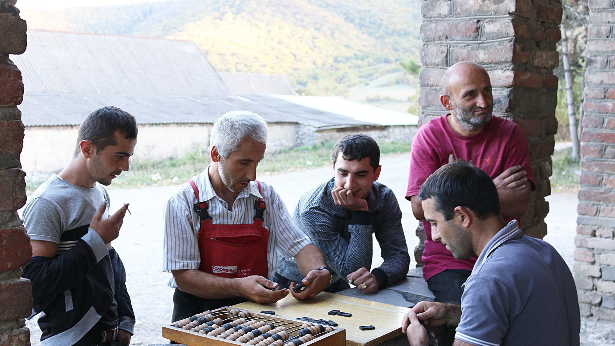 تصویر گروهی از مردان را نشان می‌دهد که در یک محیط روستایی گرد هم آمده‌اند. در مرکز تصویر، مردی با موهای سفید و لباس‌های کارگاهی به نظر می‌رسد که مشغول توضیح یا نشان دادن چیزی به دیگران است. مردان دیگر به دقت به او گوش می‌دهند و در حال بررسی یک صفحه بازی هستند که تعدادی مهره در آن قرار دارد. پس‌زمینه تصویر شامل تپه‌های سرسبز و یک ساختمان قدیمی است. جو عمومی تصویر دوستانه و غیررسمی به نظر می‌رسد.