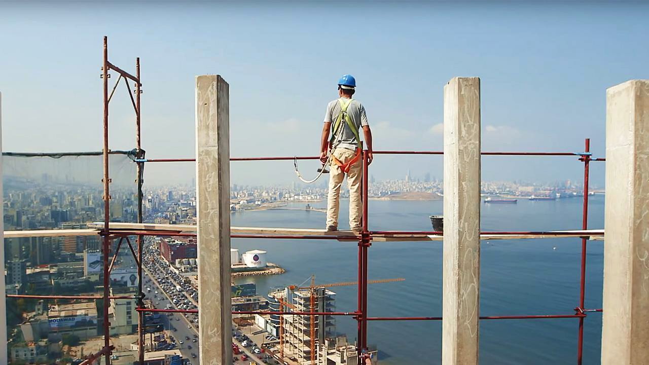 L'image montre un ouvrier de la construction se tenant sur un échafaudage, surplombant une vue panoramique sur une ville côtière. Il porte un casque de sécurité et des vêtements de travail. À l'arrière-plan, on peut voir des immeubles, une route qui longe la mer, ainsi que des bateaux sur l'eau. Le ciel est dégagé, laissant entrevoir une belle journée. La scène évoque à la fois le travail ardu des ouvriers et une magnifique perspective sur l'urbanisme et la nature environnante.