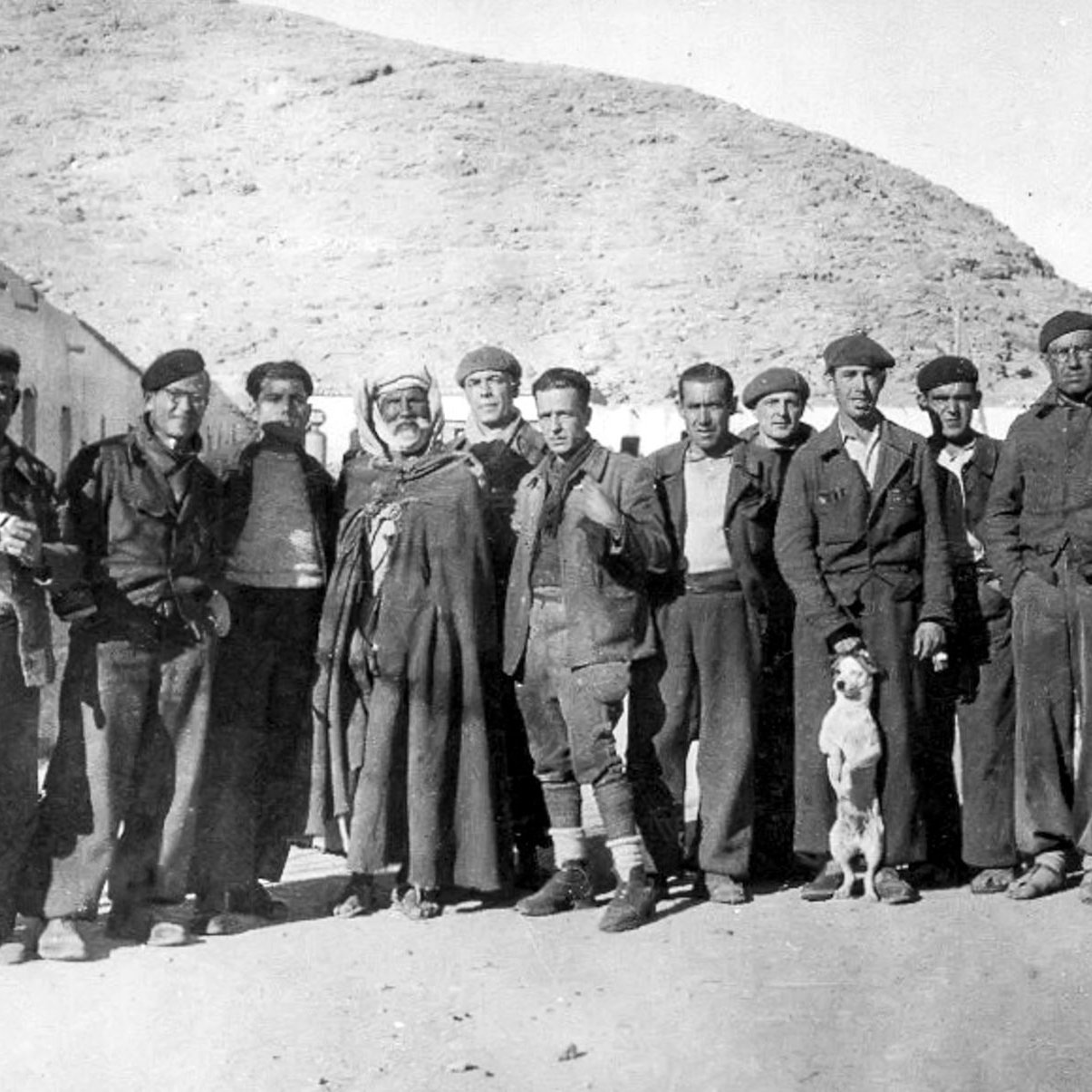 در این تصویر گروهی از مردان در یک محیط بیرونی تجمع کرده‌اند. آن‌ها به طور رسمی لباس پوشیده‌اند و در کنار هم ایستاده‌اند. در میان آن‌ها یک مرد با لباس سنتی و یک سگ دیده می‌شود. پس‌زمینه تصویر شامل تپه‌های خشک و ساختمان‌هایی به نظر می‌رسد که ویژگی‌های یک منطقه کوهستانی را نشان می‌دهد. نور تصویر قدیمی به نظر می‌رسد و به حالت سیاه و سفید است که به اثر حس نوستالژی خاصی می‌بخشد.