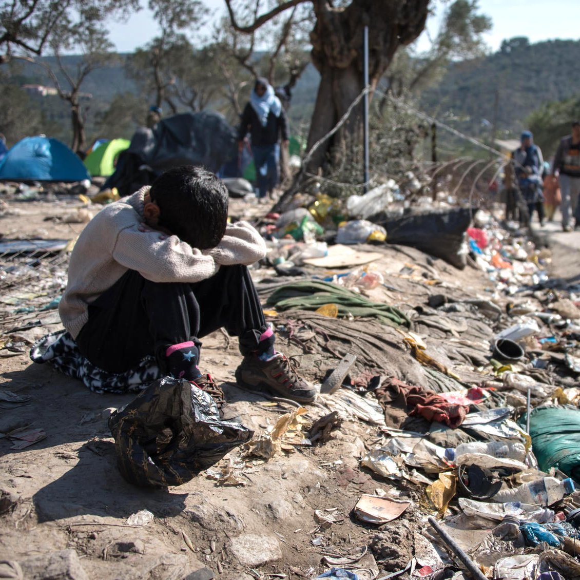 تظهر الصورة طفلاً يجلس على الأرض في ملجأ مهدم أو منطقة غير نظيفة، حيث يبدو عليه الإحباط أو الحزن، ويضع رأسه بين ركبتيه. حوله هناك أكوام من القمامة والأشياء المهملة، مما يعكس ظروف معيشة قاسية. في الخلفية، يمكن رؤية مجموعة من الأشخاص تسير في اتجاه آخر، مما يعزز الشعور بالانفصال والمعاناة. تعكس الصورة بوضوح التحديات التي يواجهها الأفراد في مثل هذه الظروف.