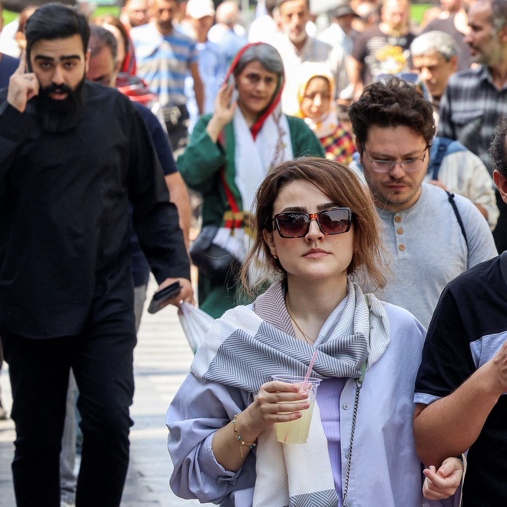 در این تصویر، گروهی از افراد در یک خیابان شلوغ در حال رفت و آمد هستند. در پیش‌زمینه، دو نفر به چشم می‌خورند که یکی از آن‌ها یک دختر با عینک بزرگ و لباس روشن است و در کنار او مردی با عینک و ریش، یک نوشیدنی در دست دارد. در پس‌زمینه، افراد دیگری نیز دیده می‌شوند که به سوی دوربین حرکت می‌کنند؛ یکی از آن‌ها مردی با ریش بلند و لباس تیره است. فضایی پرجنب‌وجوش و معمولی به نظر می‌رسد.