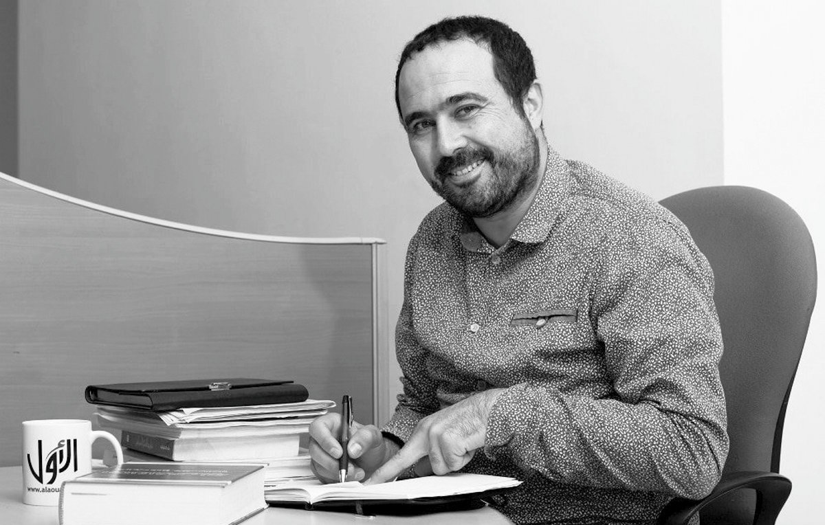 L'image montre un homme assis à un bureau, souriant et tenant un stylo dans une main tout en feuilletant un carnet ou un livre avec l'autre main. Des piles de livres sont présentes sur le bureau, ainsi qu'une tasse à café posée à côté de lui. L'environnement semble être un bureau, avec un fond minimaliste. L'image est en noir et blanc, ce qui lui donne un aspect classique.