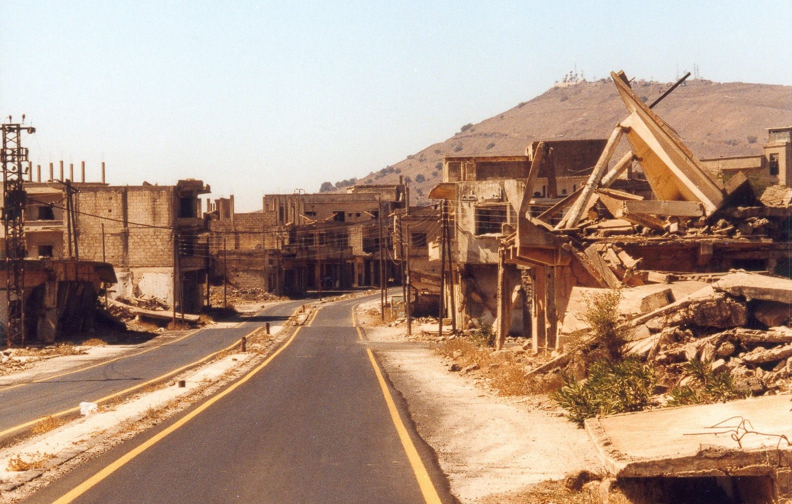 L'image montre une scène de destruction et de désolation. On peut voir une route déserte, bordée de bâtiments en ruines et de débris. Les structures semblent fortement endommagées, avec des murs effondrés et des fondations exposées. À l'arrière-plan, une colline se dresse, mais l'accent est principalement mis sur la destruction des habitations. L'atmosphère est empreinte de tristesse et d'abandon, suggérant que cet endroit a subi des conflits ou des catastrophes.