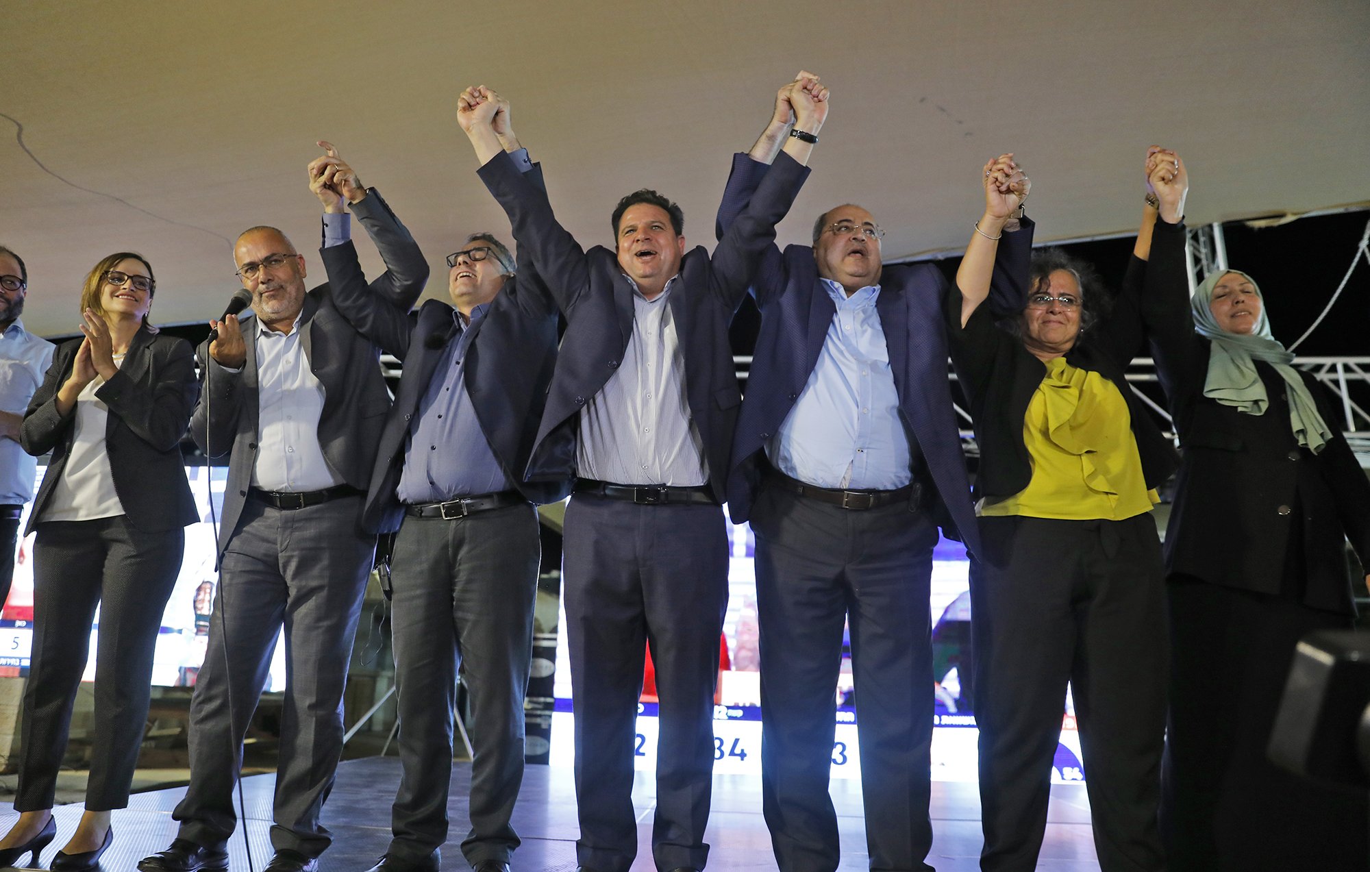 الصورة تُظهر مجموعة من الأشخاص يقفون على منصة، يرفعون أيديهم معًا في حالة من الاحتفال أو التأكيد. يبدو أنهم في مناسبة سياسية أو تجمع جماهيري، حيث تظهر خلفهم شاشات تعرض معلومات أو نتائج. يرتدي المشاركون ملابس رسمية، ويتمتعون بتعبيرات وجه تعكس الحماس والفرح.