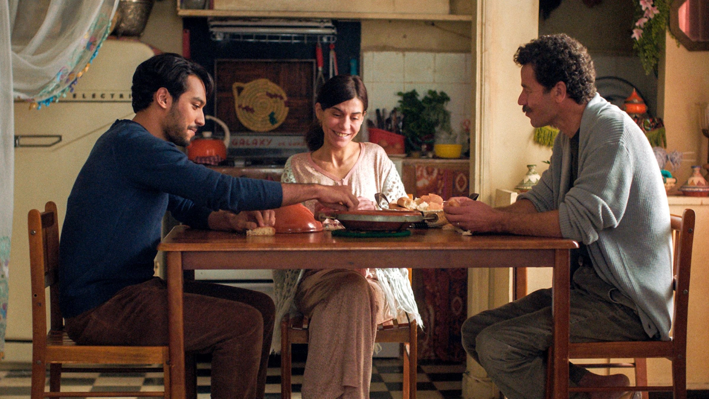 L'image montre trois personnes assises autour d'une table dans une cuisine chaleureuse et accueillante. Il y a un homme à gauche, habillé d'un pull bleu, qui sert de la nourriture à une femme souriante au centre, vêtue d'une blouse claire. À droite, un autre homme, portant un pull gris, semble également engagé dans la conversation. La cuisine est décorée de façon colorée, avec des objets et des ustensiles visibles en arrière-plan, créant une atmosphère conviviale et familiale.
