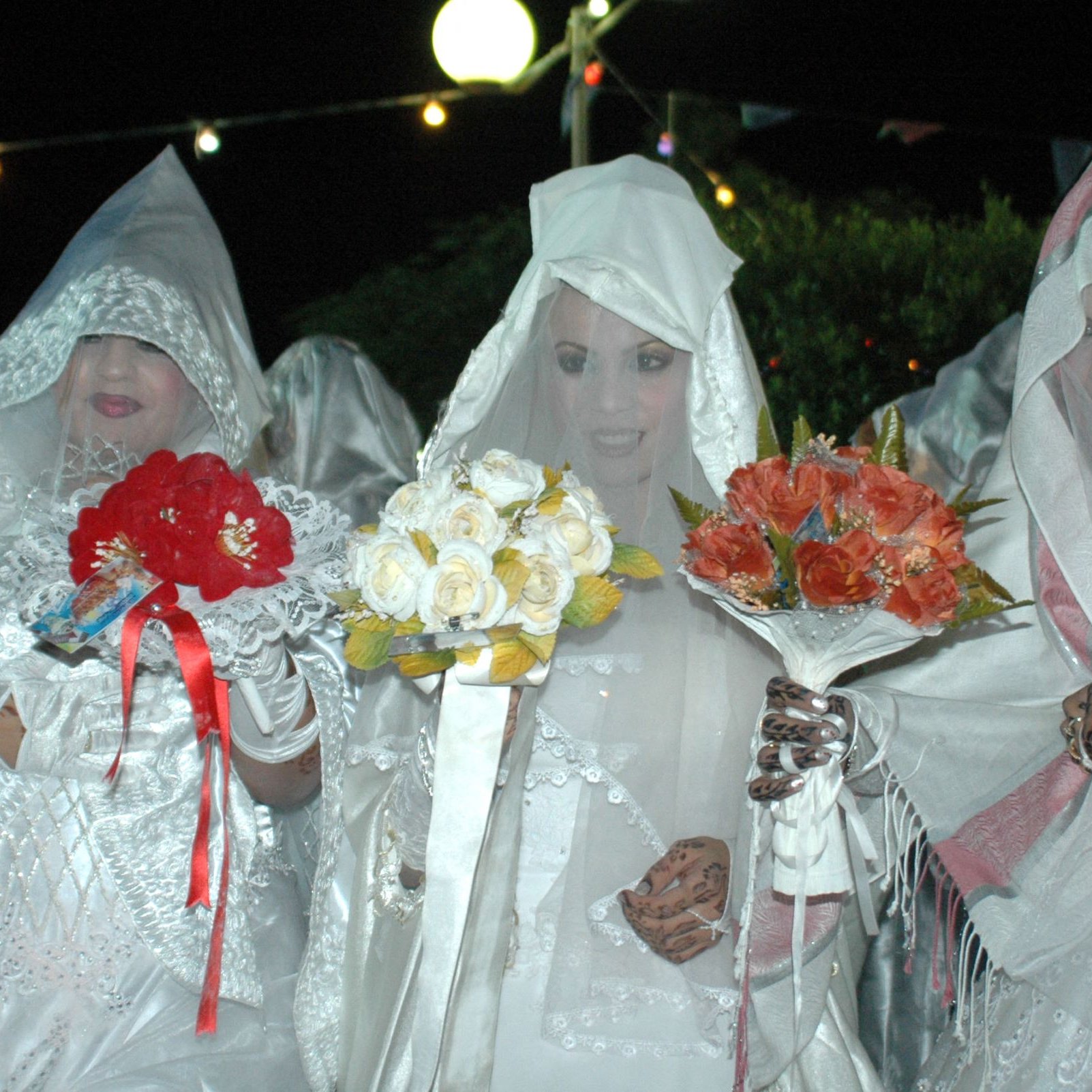L'image montre un groupe de femmes vêtues de robes blanches et de voiles, se tenant ensemble dans une ambiance nocturne. Elles tiennent chacune des bouquets de fleurs, composés de roses et d'autres fleurs colorées. L'éclairage doux crée une atmosphère festive, et des guirlandes lumineuses sont visibles en arrière-plan. Leurs visages sont en partie cachés par les voiles, ajoutant une aura de mystère à la scène.