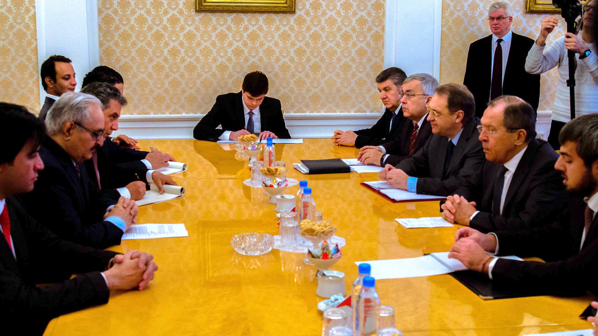 L'image présente une réunion autour d'une grande table ovale en bois. Plusieurs hommes sont assis des deux côtés, en tenue formelle, semblant être en discussion ou en train de négocier. Au centre de la table, il y a des documents et des canapés, ainsi qu'une assiette de collations. En arrière-plan, on peut voir des personnes observant la réunion, ce qui indique que c'est un moment important. L'atmosphère semble sérieuse et concentrée.