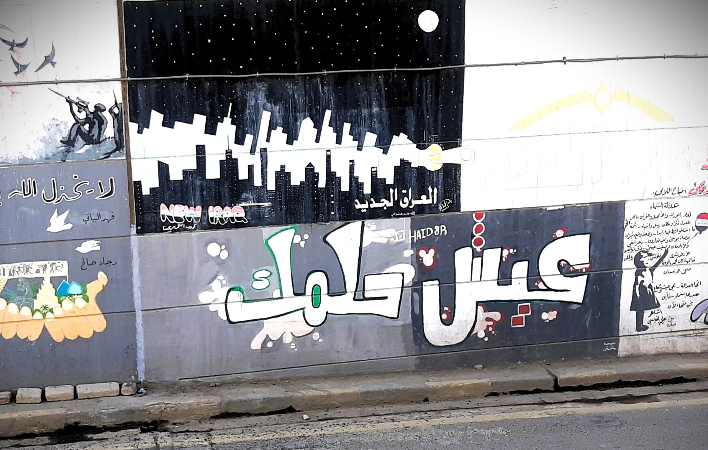 L'immagine mostra un murale colorato su un muro, con diversi disegni e scritte in arabo. In primo piano, si legge "عيش حلمك" che significa "Vivi il tuo sogno". Lo sfondo presenta vari elementi artistici, tra cui silhouette di grattacieli e figure stilizzate, creando un'atmosfera urbana e vibrante. Ci sono anche elementi decorativi e simboli che riflettono un messaggio di speranza e ispirazione.
