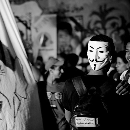 L'image montre une scène de rassemblement ou de manifestation. Un groupe de personnes est réuni, et au premier plan, une personne porte un masque stylisé, souvent associé au mouvement Anonymous. En arrière-plan, on peut voir une grande bannière avec des illustrations ou des visages, probablement liés à un message politique. L'atmosphère semble dynamique, avec des expressions de joie ou de solidarité parmi les participants. Le tout est en noir et blanc, ce qui accentue l'intensité de la scène.