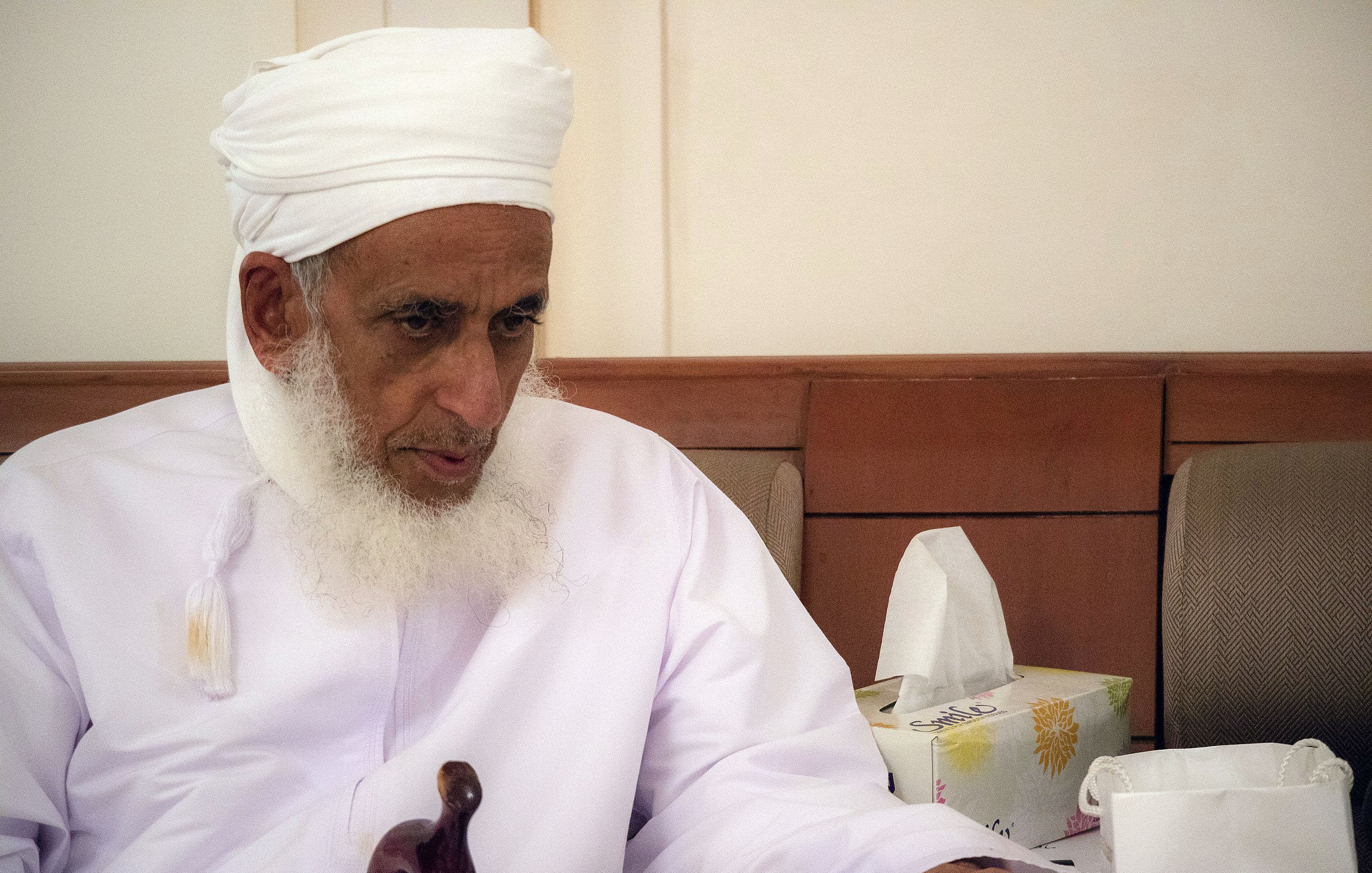 تظهر في الصورة رجل مسن يجلس في مكان مغلق. يرتدي روبًا تقليديًا أبيض مع عمامة بيضاء، ويبدو عليه الوقار والحكمة. لديه لحية بيضاء طويلة، وينظر إلى شيء ما أمامه بتركيز. بجانبه توجد علبة مناديل ورقية، مما يوحي بأنها بيئة اجتماعية أو مريحة. تعكس تعابيره احترامه وتفكيره العميق.