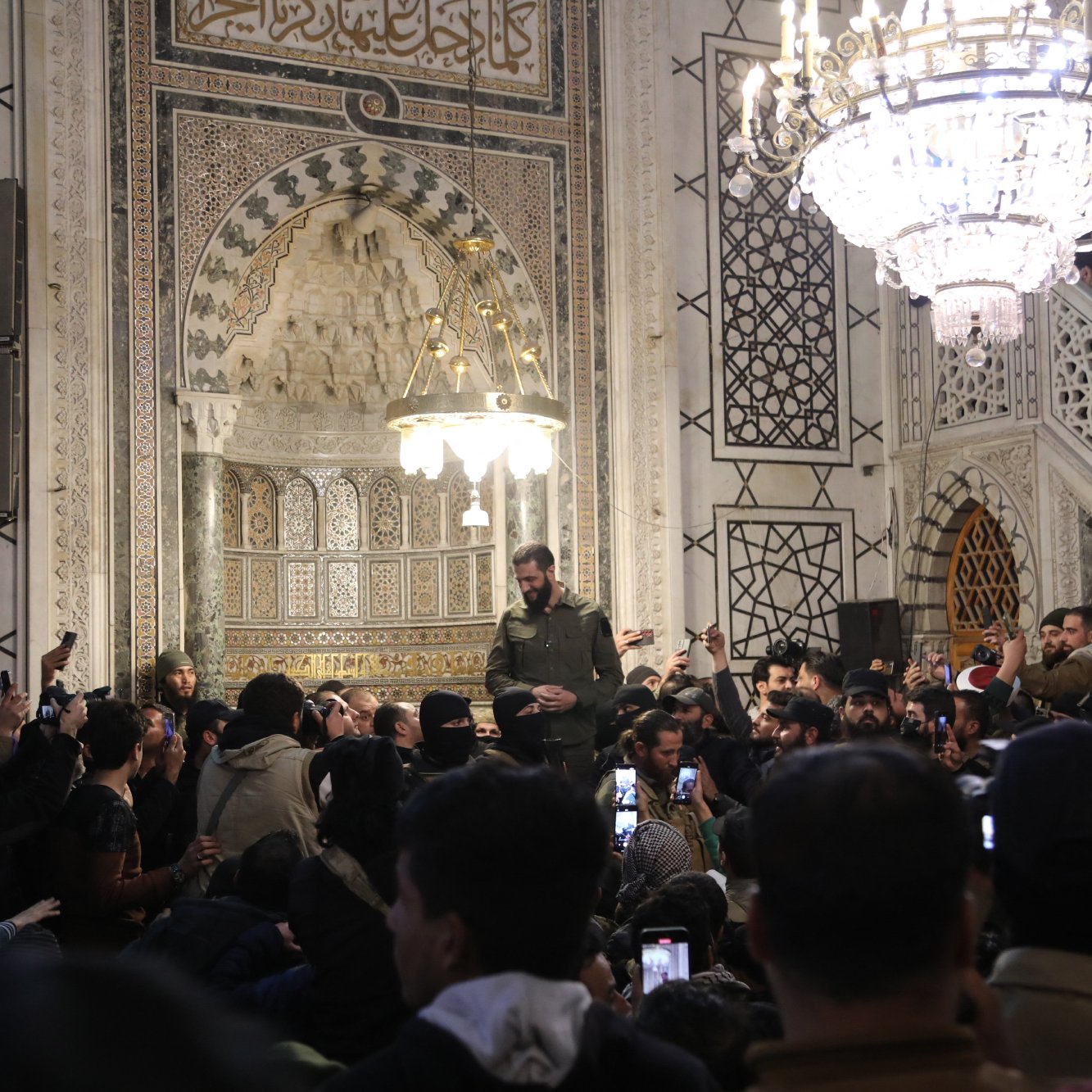 تظهر الصورة مجموعة من الأشخاص في مكان داخلي مزين بتفاصيل معمارية جميلة. هناك شخص يقف في الوسط، يبدو أنه يتحدث أو يلقي كلمة، بينما يحيط به حشد من المشاركين الذين يلتقطون الصور بأجهزتهم المحمولة. الإضاءة في المكان تتضمن ثريا كبيرة تعكس طابعًا تاريخيًا وثقافيًا، مع وجود عناصر زخرفية معمارية مميزة على الجدران. يبدو أن الحدث مهم وجذب انتباه الحضور بشكل كبير.