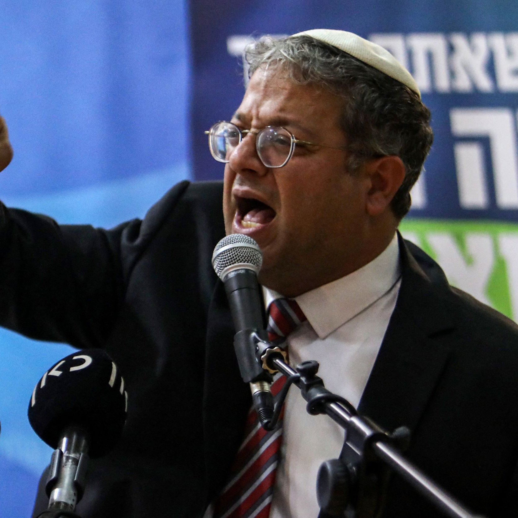 L'image montre un homme portant des lunettes et une kippa, en train de s'exprimer devant un micro. Il semble passionné par ses propos, faisant un geste avec son bras droit tendu et son poing fermé. Il porte un costume sombre avec une cravate rayée. Devant lui, plusieurs micros de différentes chaînes de presse sont visibles. L'arrière-plan montre une bannière avec du texte en hébreu.