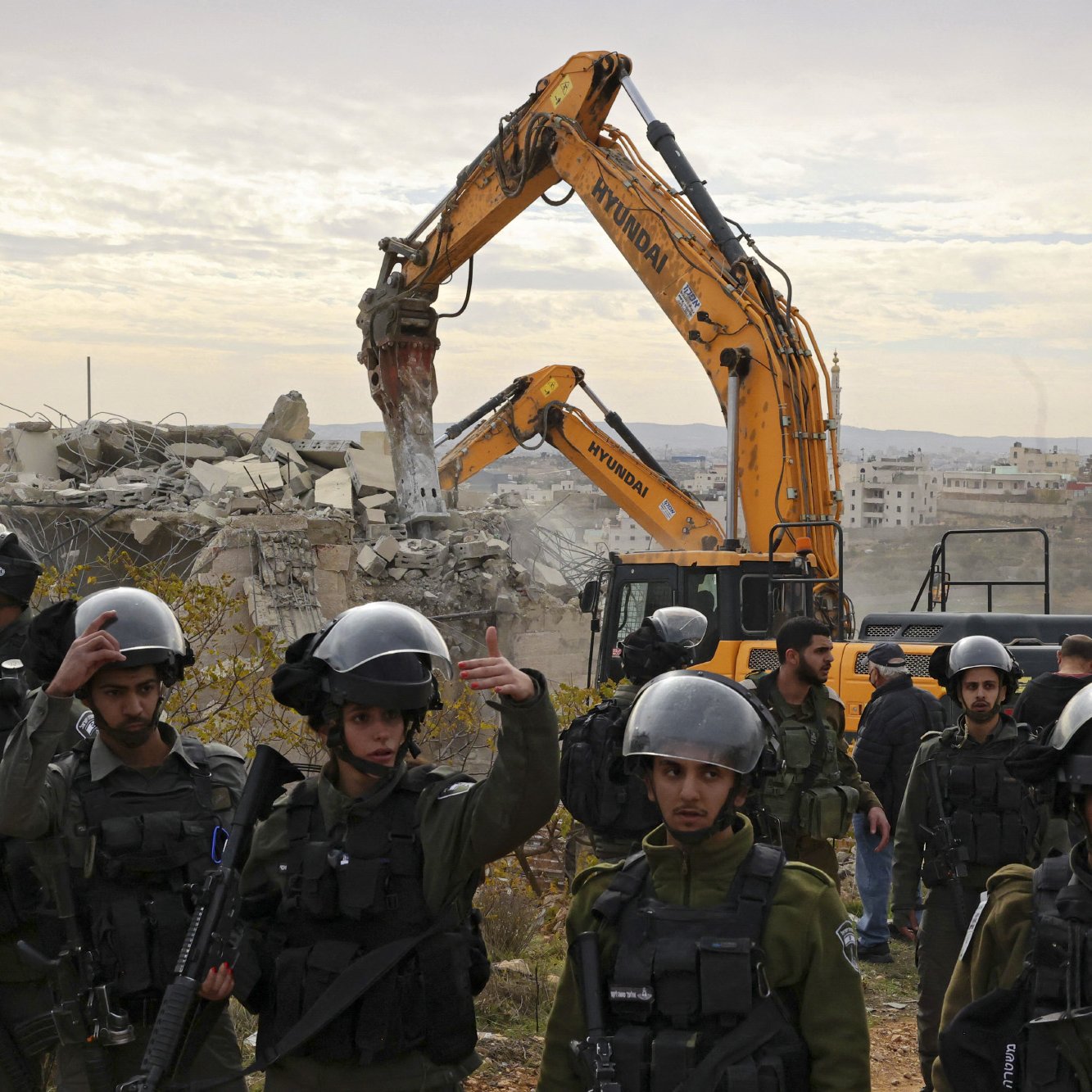 L'image montre une scène de destruction, où un bulldozer démolit un bâtiment en arrière-plan. Au premier plan, un groupe de soldats portant des casques et des équipements de protection surveille la situation. Ils semblent être en alerte, avec des armes en main. L'environnement est marqué par des décombres et un paysage urbain, suggérant un contexte de conflit. Le ciel est nuageux, ajoutant une atmosphère somber à la scène.