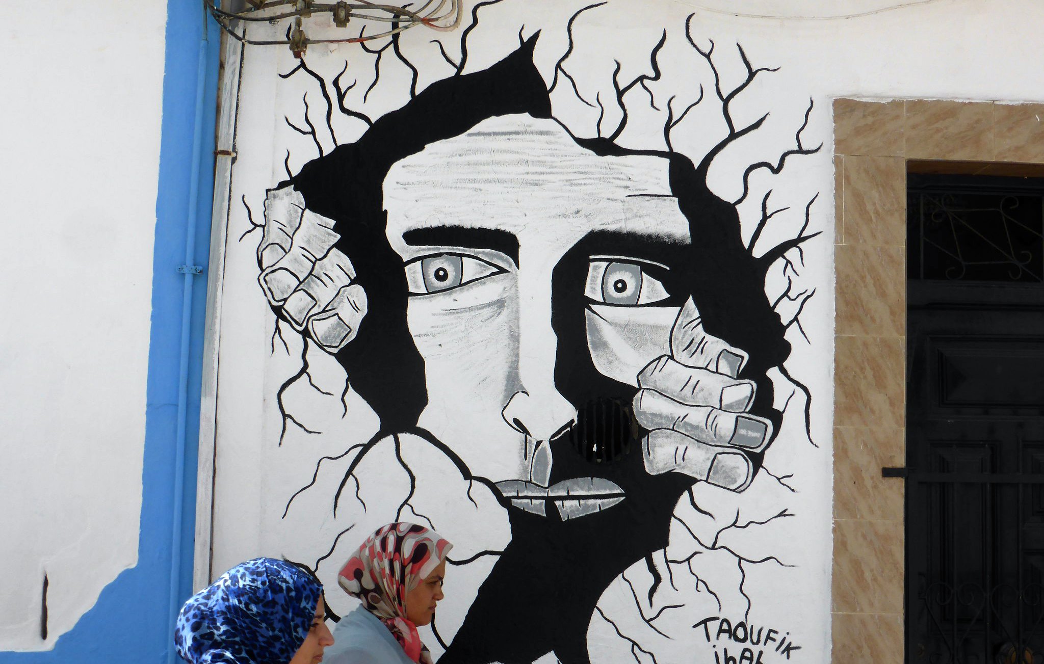 L'image montre une fresque murale en noir et blanc représentant un visage humain au milieu d'une apparente fissure dans le mur. Le visage a des traits marqués et semble exprimer une émotion intense. Des mains émergent des fissures, comme si elles tentaient de sortir. Au premier plan, deux femmes en hijab marchent, apportant une touche de couleur et de vie à la scène. Le tout crée un contraste entre l'art mural et la réalité environnante.