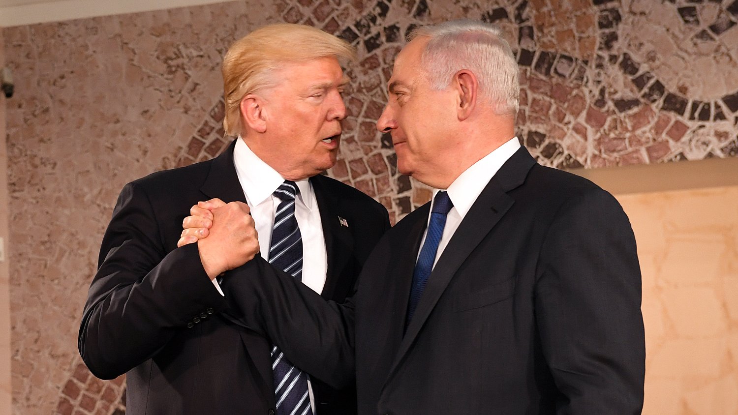 L'image montre deux hommes en train de se serrer la main avec des expressions sérieuses. Ils sont habillés en costume, l'un portant une cravate rayée et l'autre une cravate unie. L'arrière-plan possède des motifs décoratifs. Cette scène semble se dérouler lors d'un événement officiel ou d'une rencontre diplomatique.