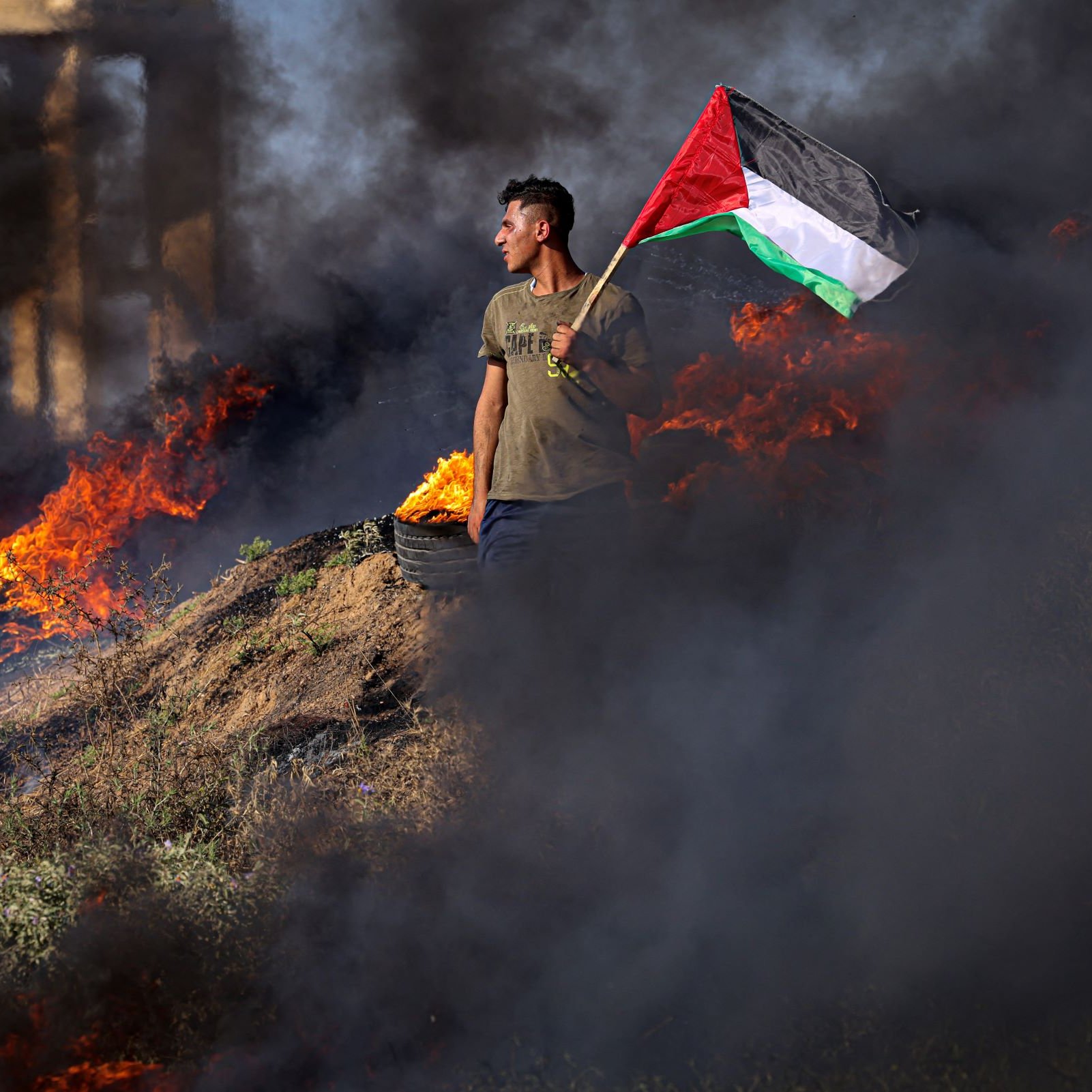 تظهر الصورة شابًا يحمل علم فلسطين، يقف وسط دخان كثيف وحرائق مستعرة. يبدو أن الخلفية تعكس حالة من التوتر أو الصراع، حيث النيران تشتعل بالقرب منه. يرتدي الشاب زيًا عاديًا، مما يشير إلى أنه في أوضاع ميدانية. تعكس الصورة مشاعر العزة والانتماء الوطني.