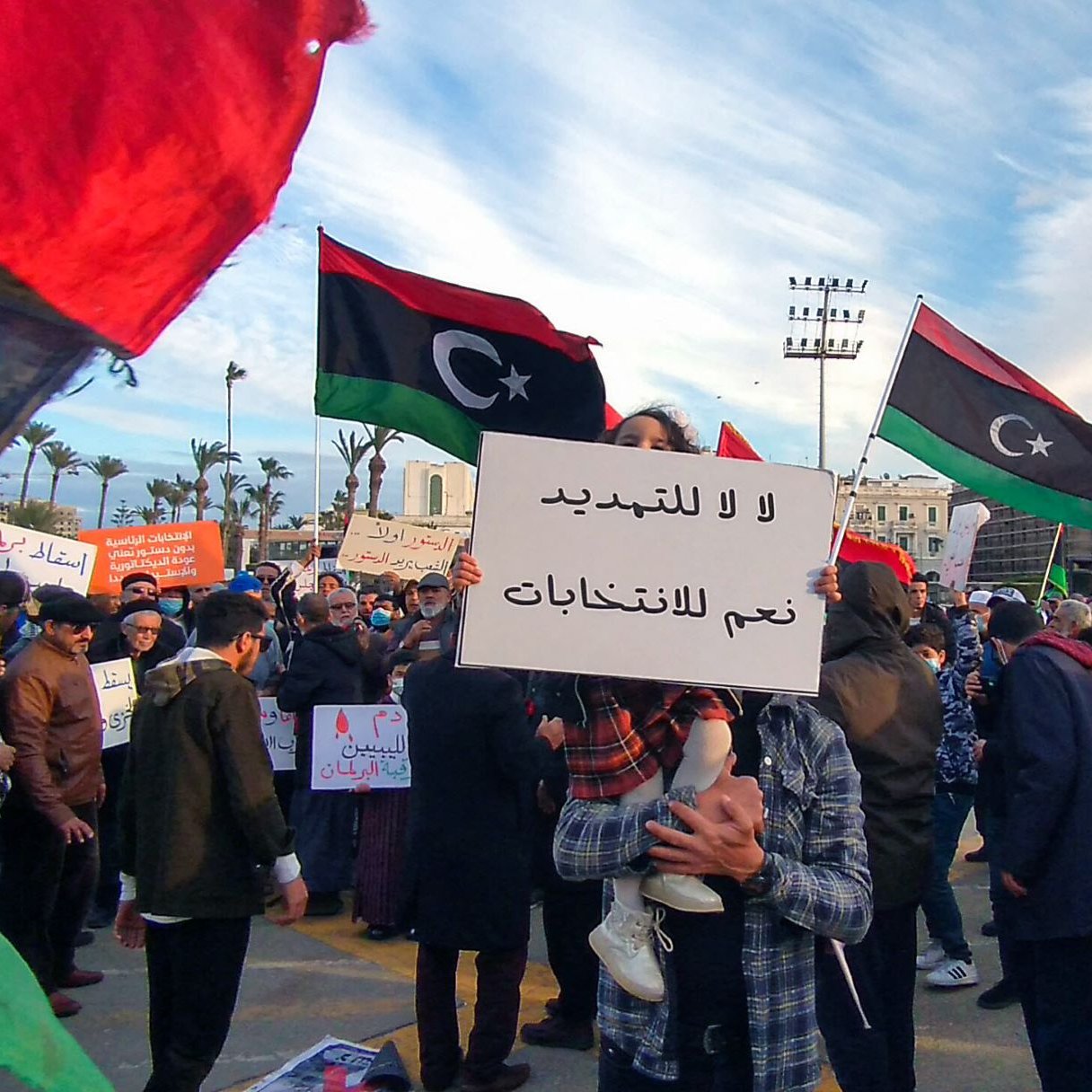 L'immagine ritrae una manifestazione vivace, con molte persone in piedi in un'area aperta. Alcuni manifestanti tengono cartelli, tra cui uno che dice "لا للتعديل" (No alle modifiche) e "نعم للانتخابات" (Sì alle elezioni). Sono visibili anche bandiere libiche, con i loro colori distintivi di rosso, nero e verde. Lo sfondo presenta alberi e torri, e c'è un'atmosfera di protesta pacifica, con i partecipanti che sembrano impegnati a esprimere le loro opinioni su questioni politiche.