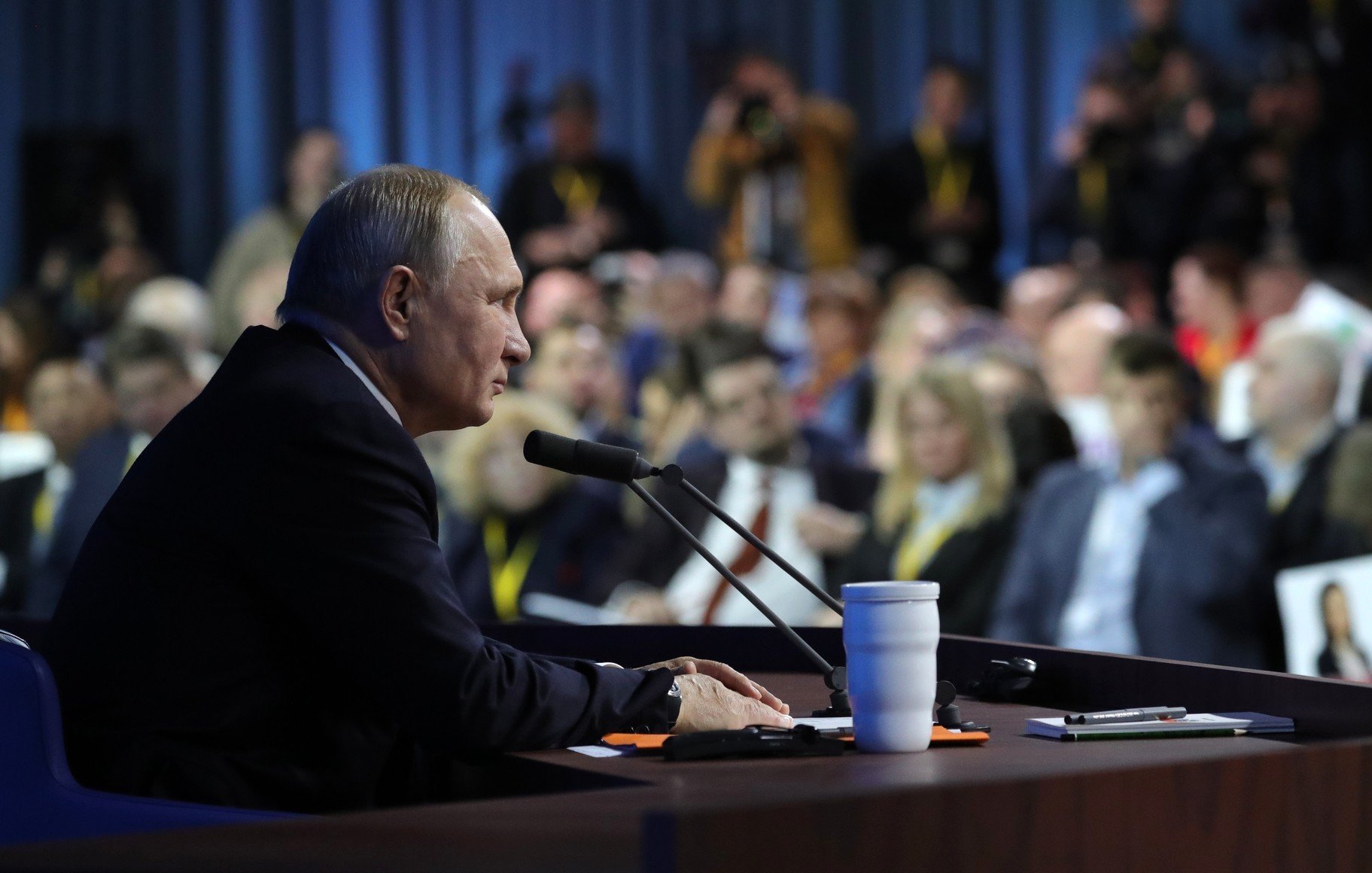 در این تصویر، ولادیمیر پوتین، رئیس‌جمهور روسیه، در حال سخنرانی یا پاسخ به سؤالات در یک کنفرانس مطبوعاتی به نظر می‌رسد. او در حال نشستن در کنار یک میز است که میکروفون دارد و در مقابل او تعدادی از خبرنگاران و حضار به نظر می‌رسند. محیط اطراف کمی تاریک‌تر است و تمرکز اصلی بر روی پوتین قرار دارد. تعدادی از افراد حاضر در جمع در پس‌زمینه دیده می‌شوند.