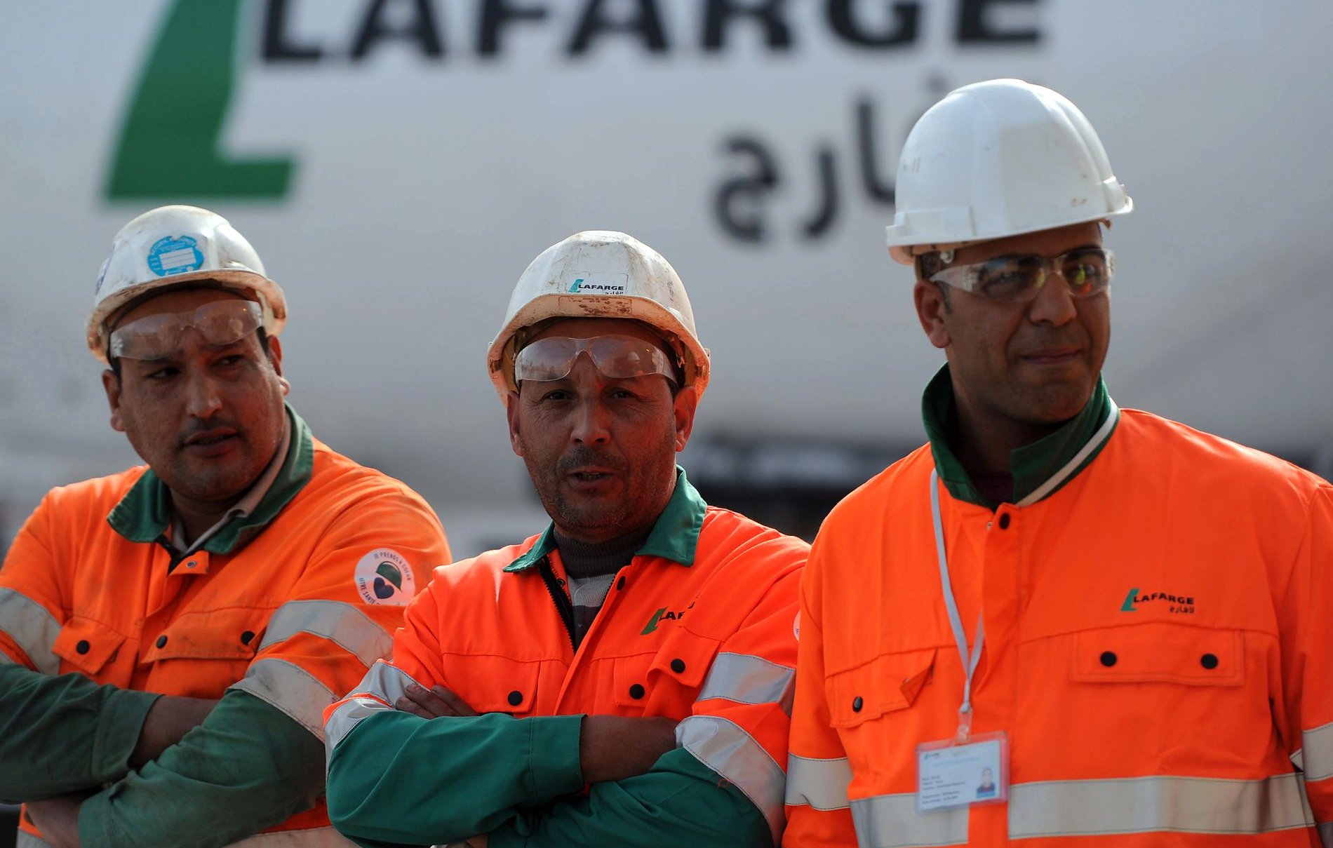 L'image montre trois ouvriers portant des vêtements de sécurité orange et des casques blancs. Ils se tiennent debout avec les bras croisés, affichant une attitude sérieuse. En arrière-plan, on peut voir un camion ou un véhicule avec le logo de Lafarge, une entreprise connue dans le secteur des matériaux de construction. L'environnement semble être un chantier ou une zone industrielle.