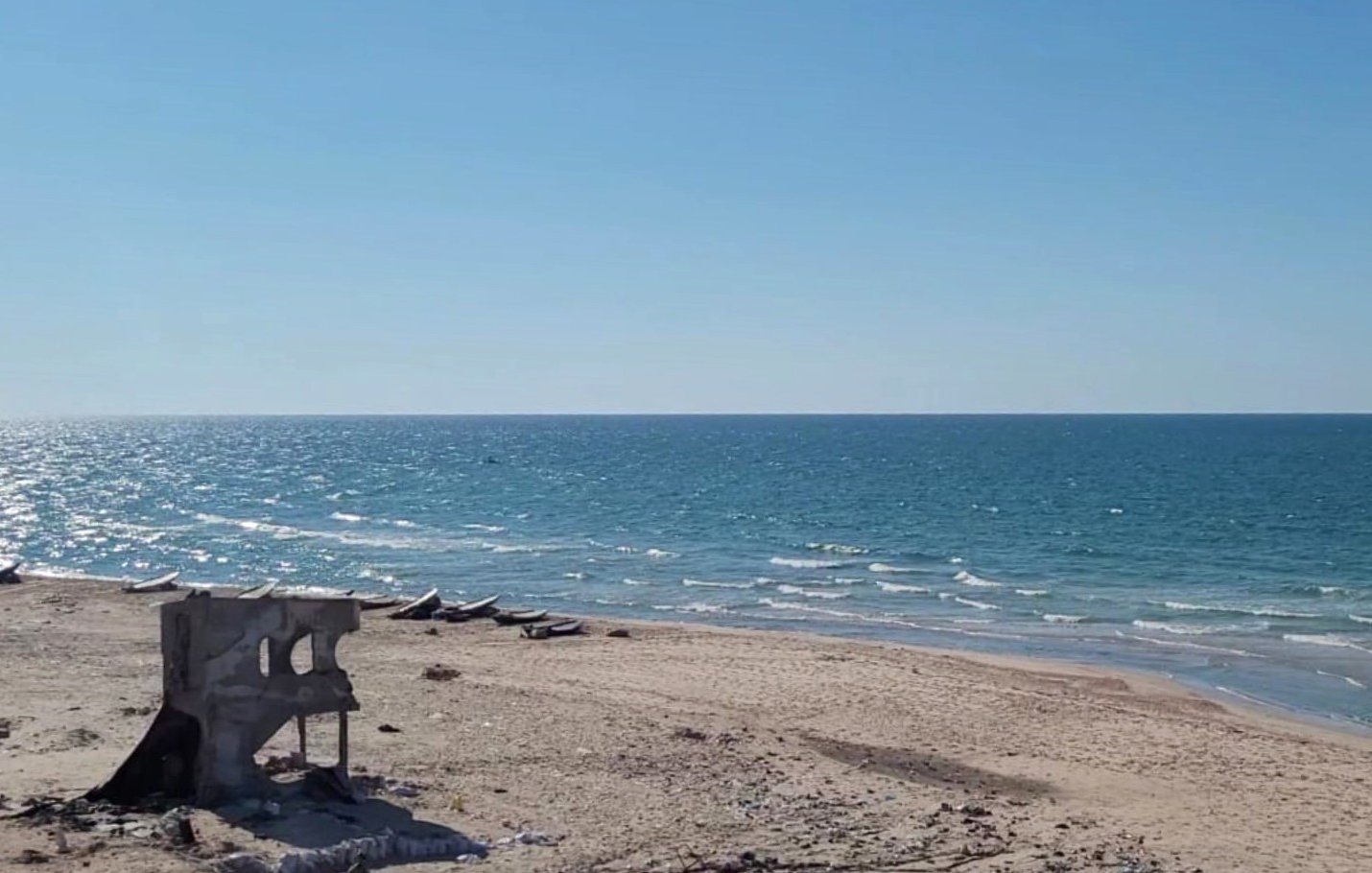 L'image montre une plage sous un ciel bleu clair, avec une mer d'un bleu éclatant qui s'étend à perte de vue. Le rivage est bordé de sable fin, mais des débris, comme une structure en béton en ruine, sont visibles sur la plage, ajoutant un contraste à la beauté naturelle du paysage. Au loin, on peut apercevoir quelques petites embarcations sur l'eau, suggérant une ambiance tranquille et isolée.