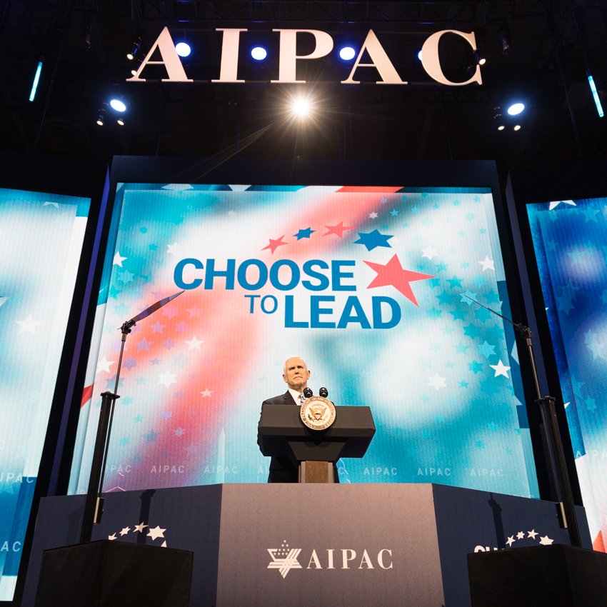L'image montre un homme sur scène, probablement un orateur, devant un public. Il se tient derrière un pupitre avec le logo d'AIPAC, une organisation pro-israélienne. En arrière-plan, il y a un grand écran affichant le slogan "CHOOSE TO LEAD" (Choisissez de diriger), entouré de motifs étoilés. L'ambiance semble formelle et dédiée à un discours important, probablement lors d'une conférence ou d'un événement lié à AIPAC.