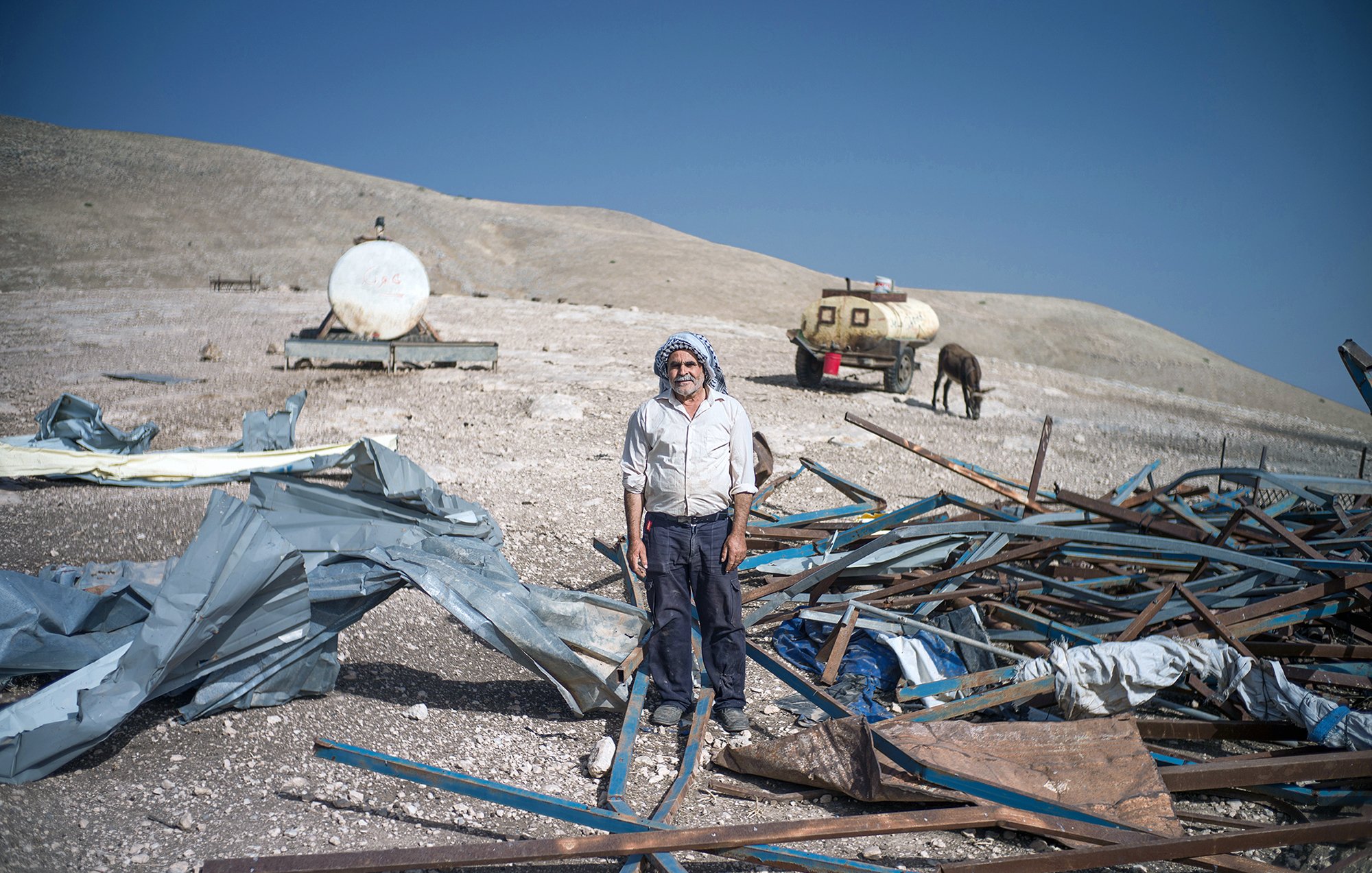 L'image montre un homme debout au milieu d'un terrain désolé. Il est entouré de débris, comprenant des structures métalliques et des morceaux de toile. En arrière-plan, on peut voir des réservoirs ou des citernes, ainsi qu'un cheval. Le paysage semble aride, avec des collines en arrière-plan sous un ciel bleu. L'homme porte une tenue simple et dégage une impression de résilience dans un environnement difficile.