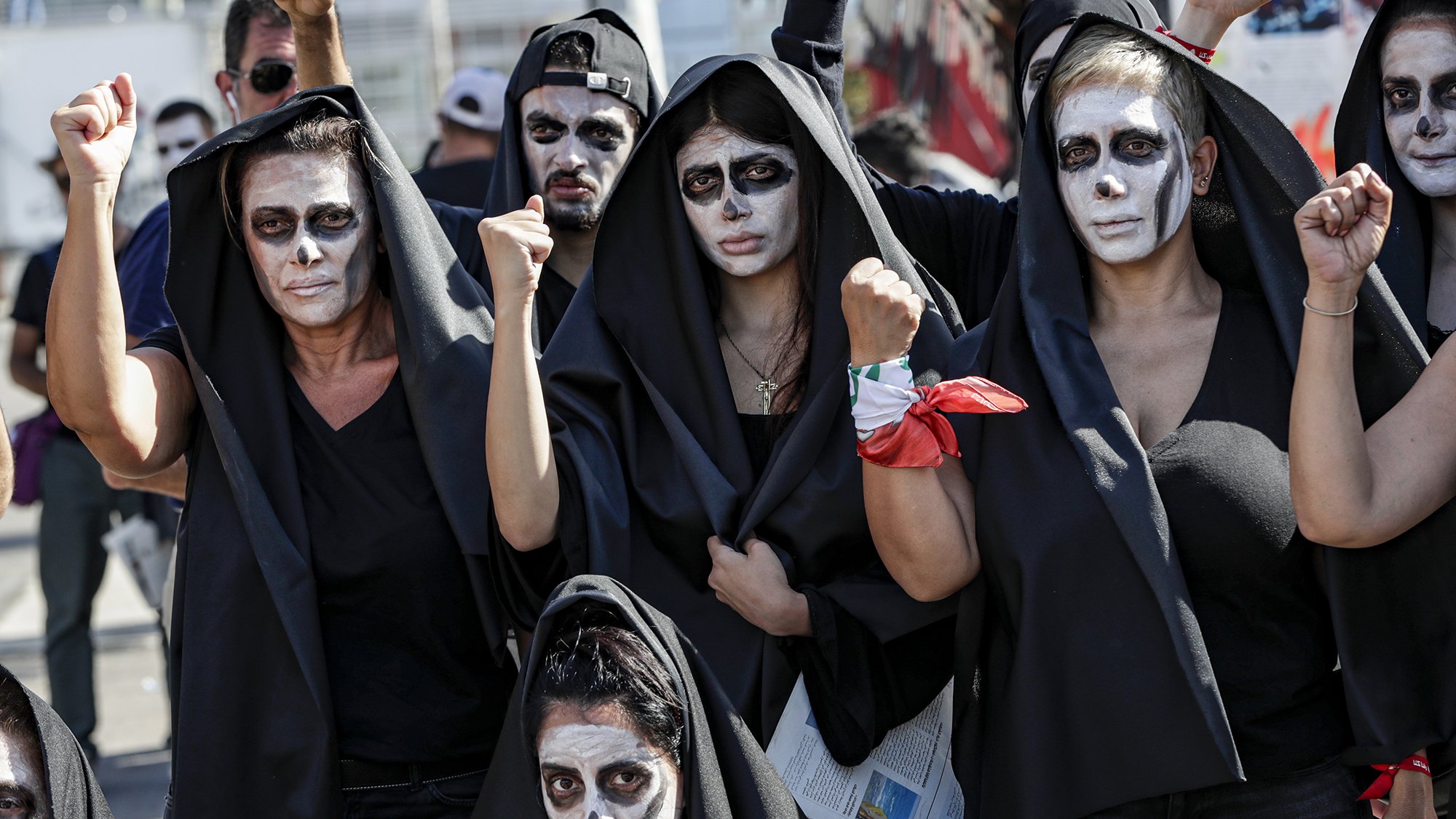 L'image montre un groupe de personnes, principalement des femmes, portant des vêtements noirs et des capes. Leur visage est peint en blanc avec des accents noirs, et elles levèrent les poings de manière déterminée. L'atmosphère semble être celle d'une manifestation ou d'une protestation, avec une ambiance de solidarité et de revendication. Certains participants portent un foulard ou un accessoire rouge. L'ensemble dégage un sentiment fort de résistance et d'engagement.