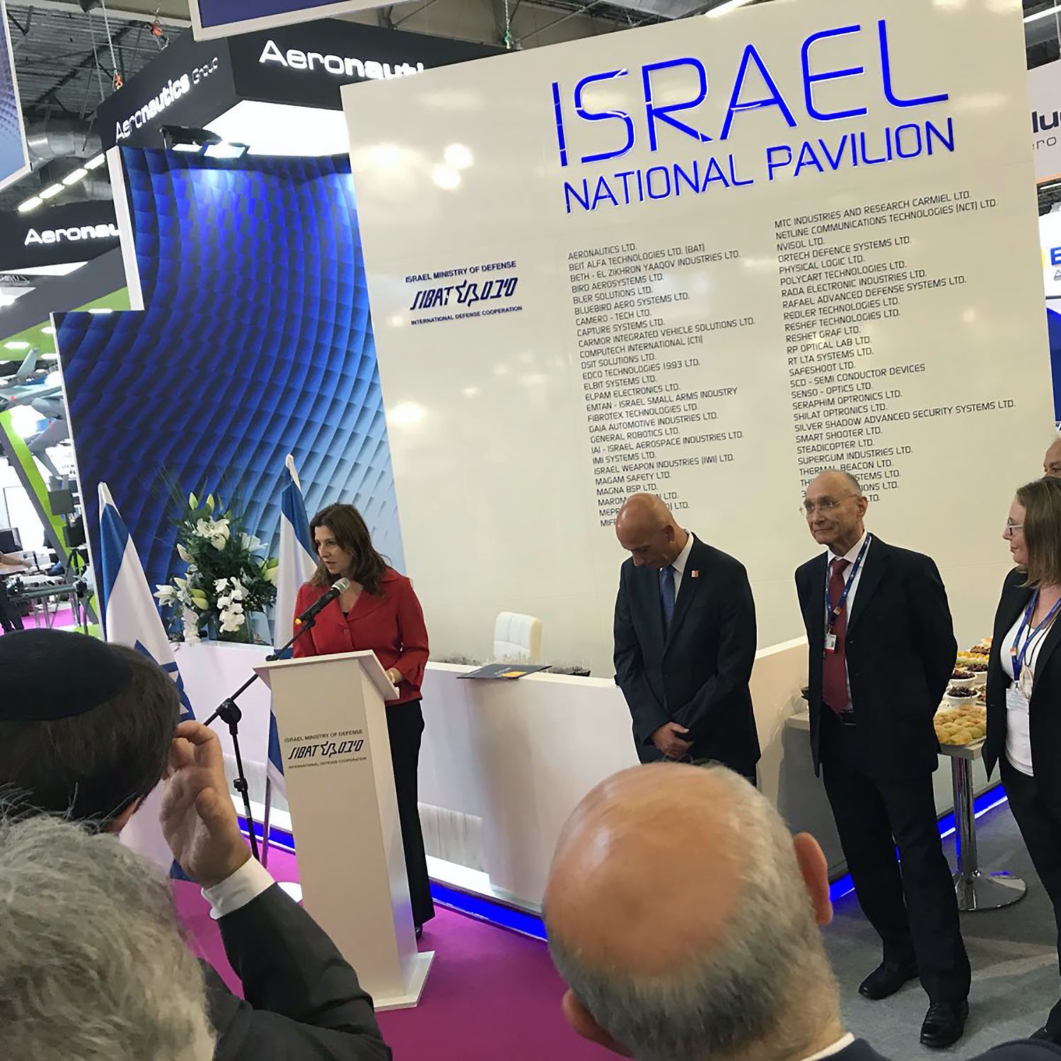 The image shows a gathering at the Israel National Pavilion at an event or exhibition. A woman is speaking at a podium, while several individuals stand beside her, including a man in a suit and others who appear to be attentive to her speech. The background features a large display with the words “ISRAEL NATIONAL PAVILION” and various company names listed. The atmosphere seems formal, with an audience present, possibly indicating a business or governmental event.